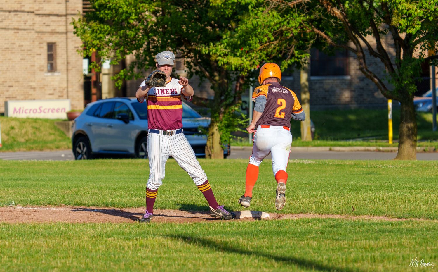 Two youth baseball players on field, one fielder catching ball at base while runner in orange uniform approaches.