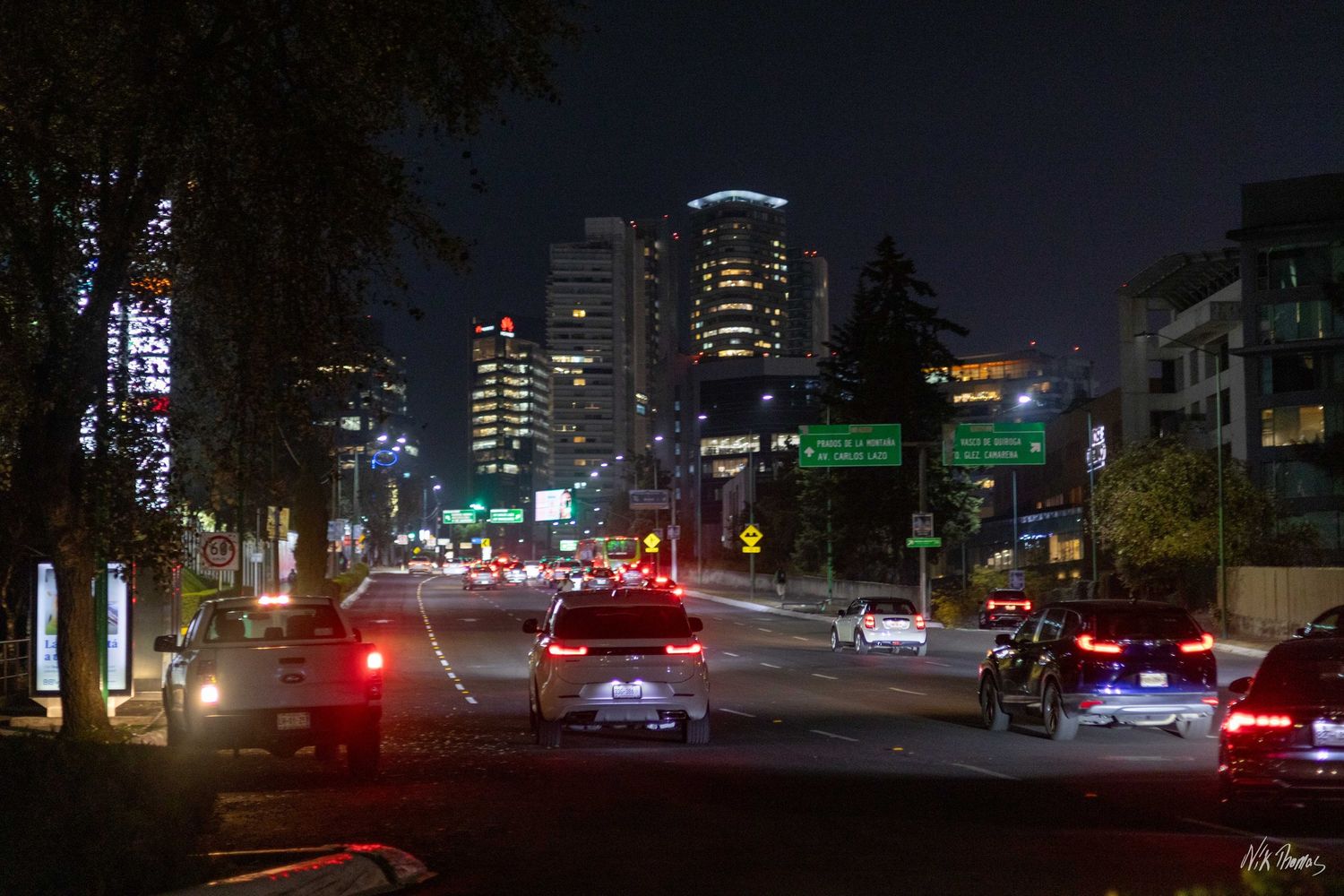Nighttime city street with cars and glowing red taillights, illuminated skyscrapers and green traffic signs in background.