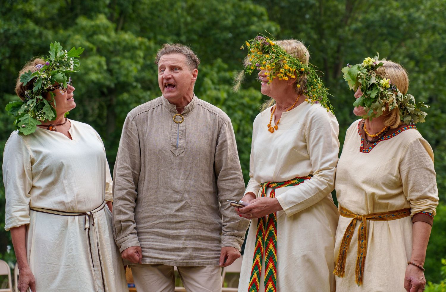 Four people in traditional folk costumes and floral wreaths singing outdoors at a cultural festival.