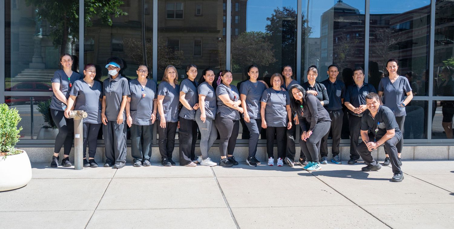 Group of healthcare workers in gray uniforms posing together outside a modern building on a sunny day.