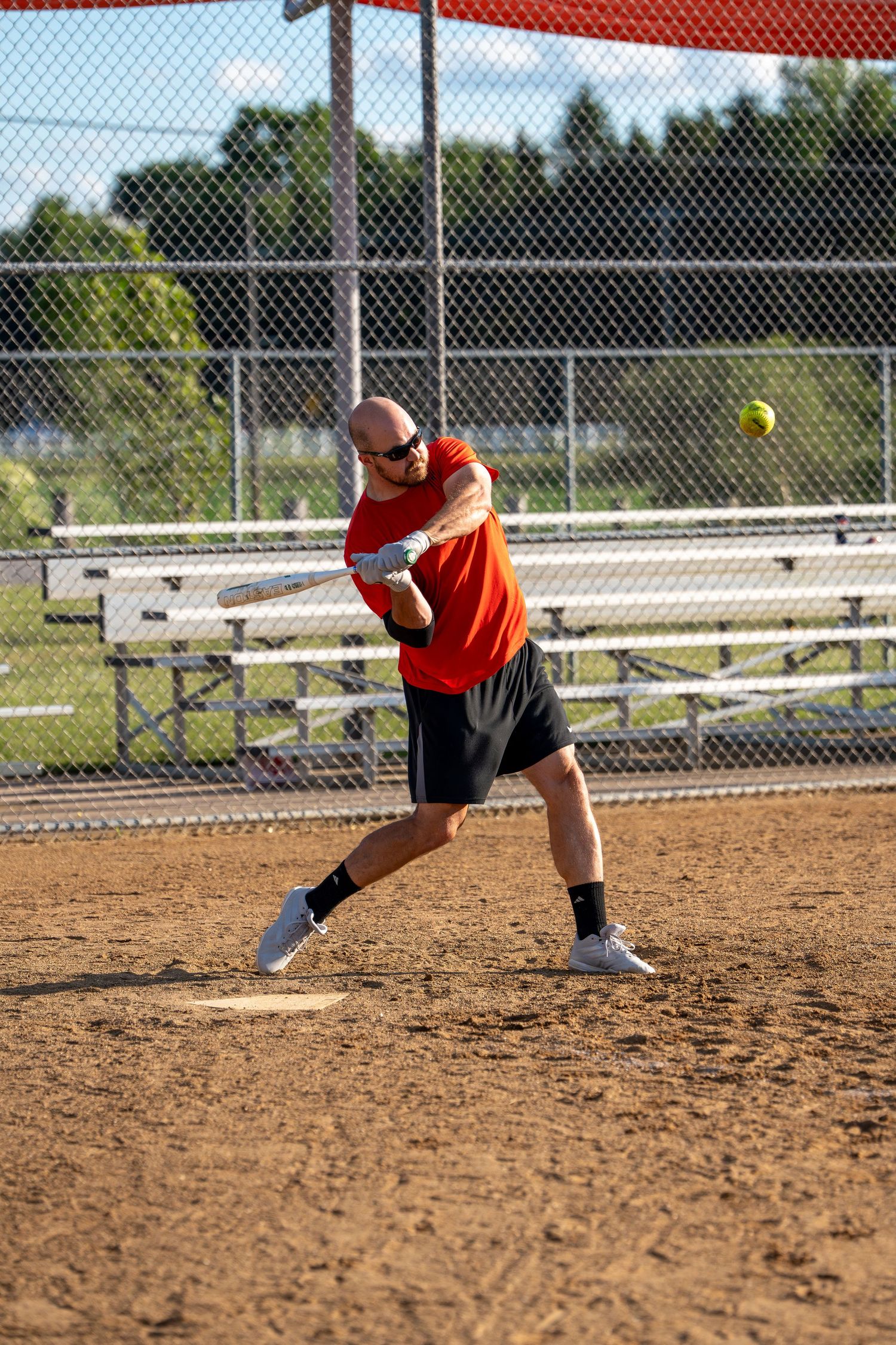 Bald man in red shirt powerfully swinging a bat at a yellow softball on a sunny outdoor dirt field.