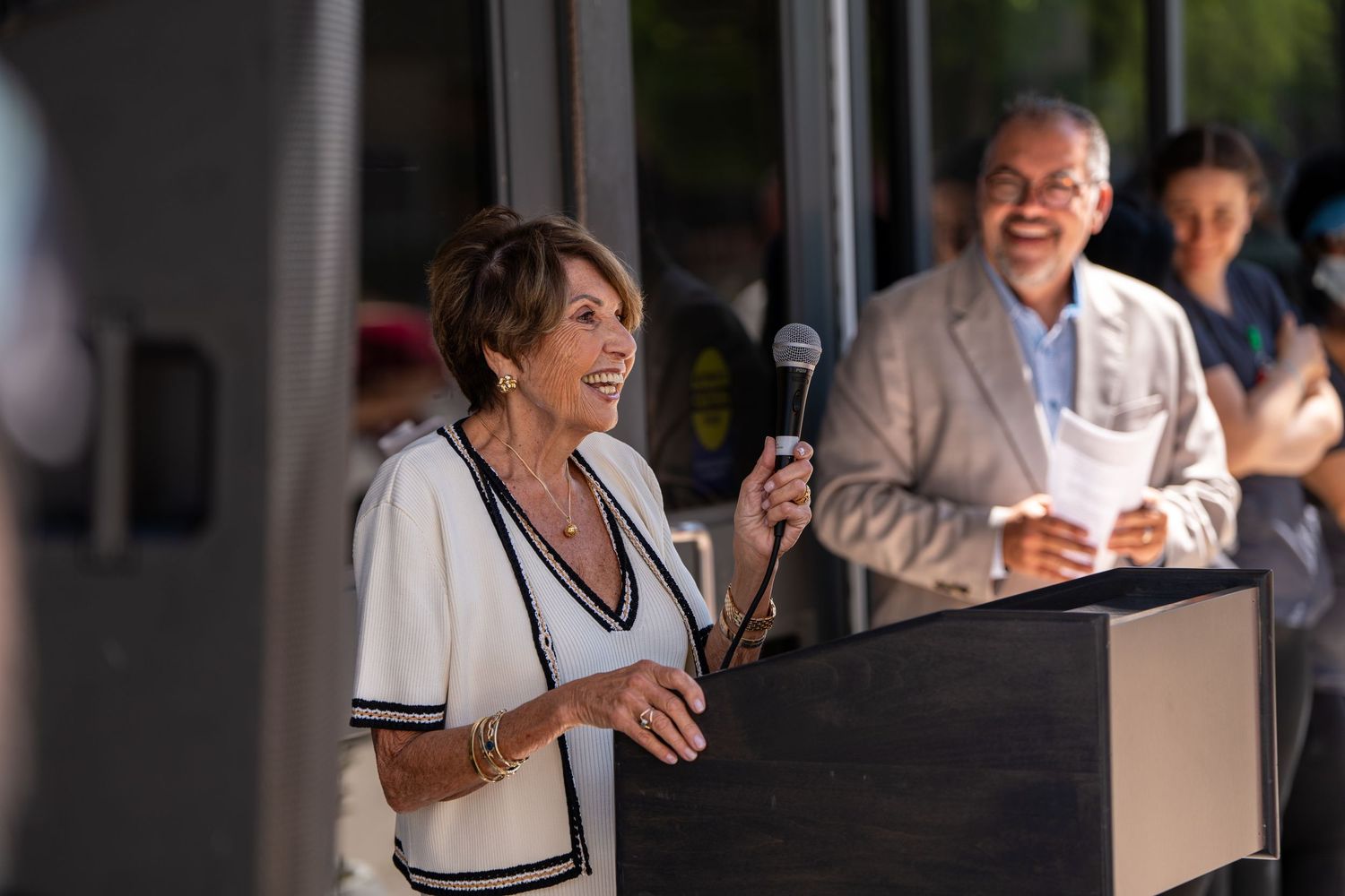 Woman in white jacket speaks at podium with microphone at outdoor event with man in suit behind her.