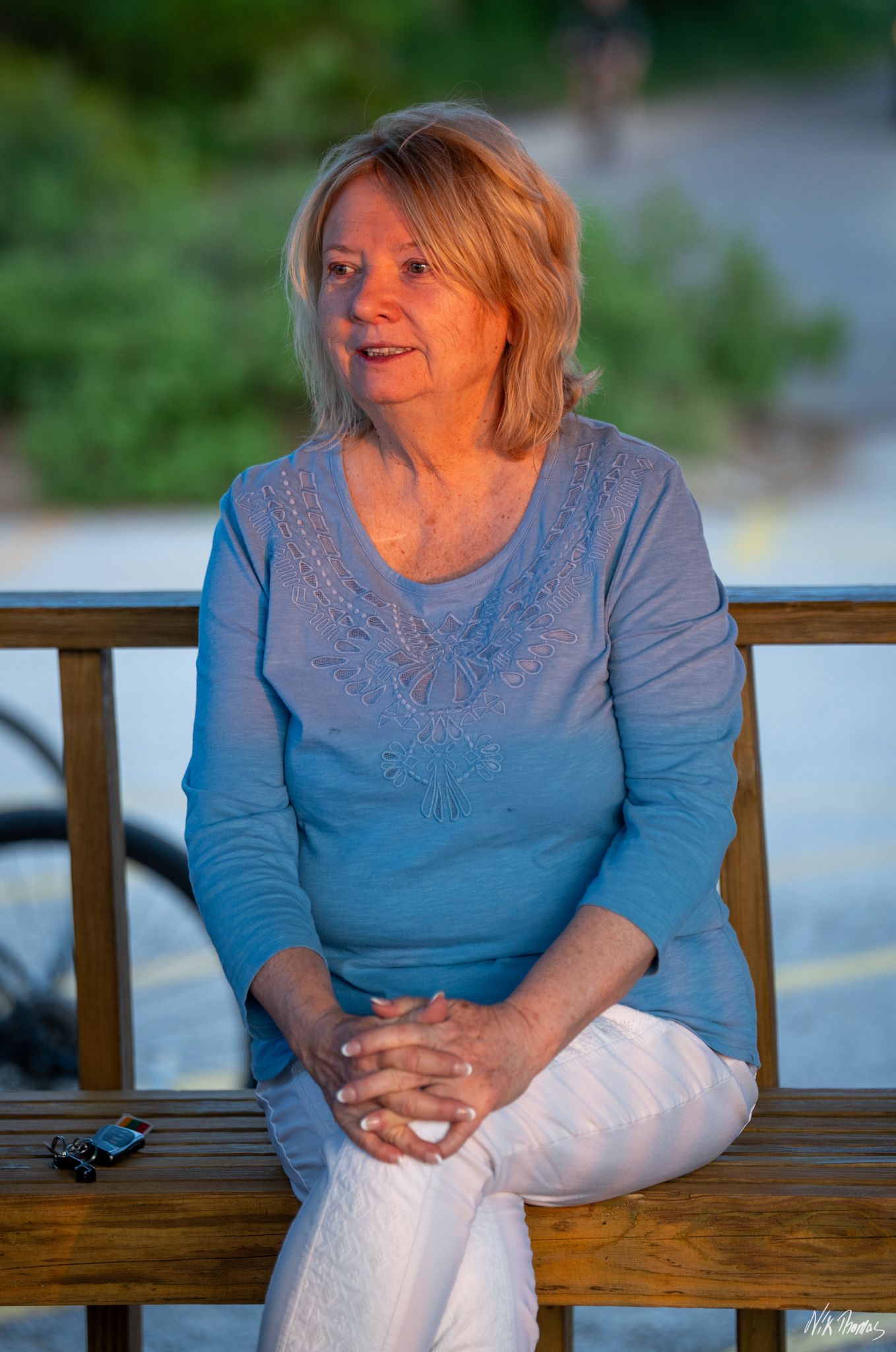 A smiling woman with blonde hair sits on a wooden bench wearing a blue top and white pants.