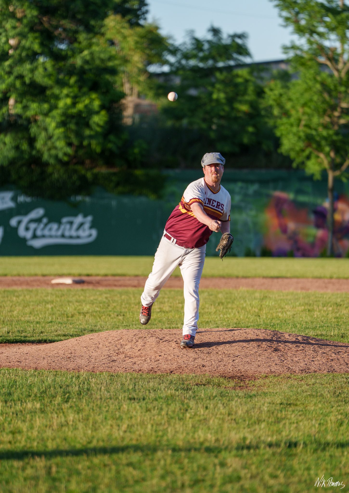 Baseball pitcher in maroon uniform mid-throw on mound with Giants sign visible in background.
