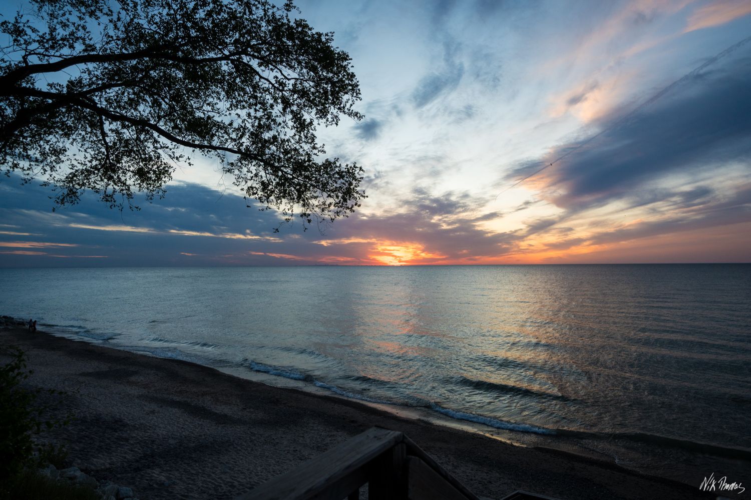Scenic lakeside sunset with vibrant orange sky, dramatic clouds, and silhouetted tree branches over calm water.
