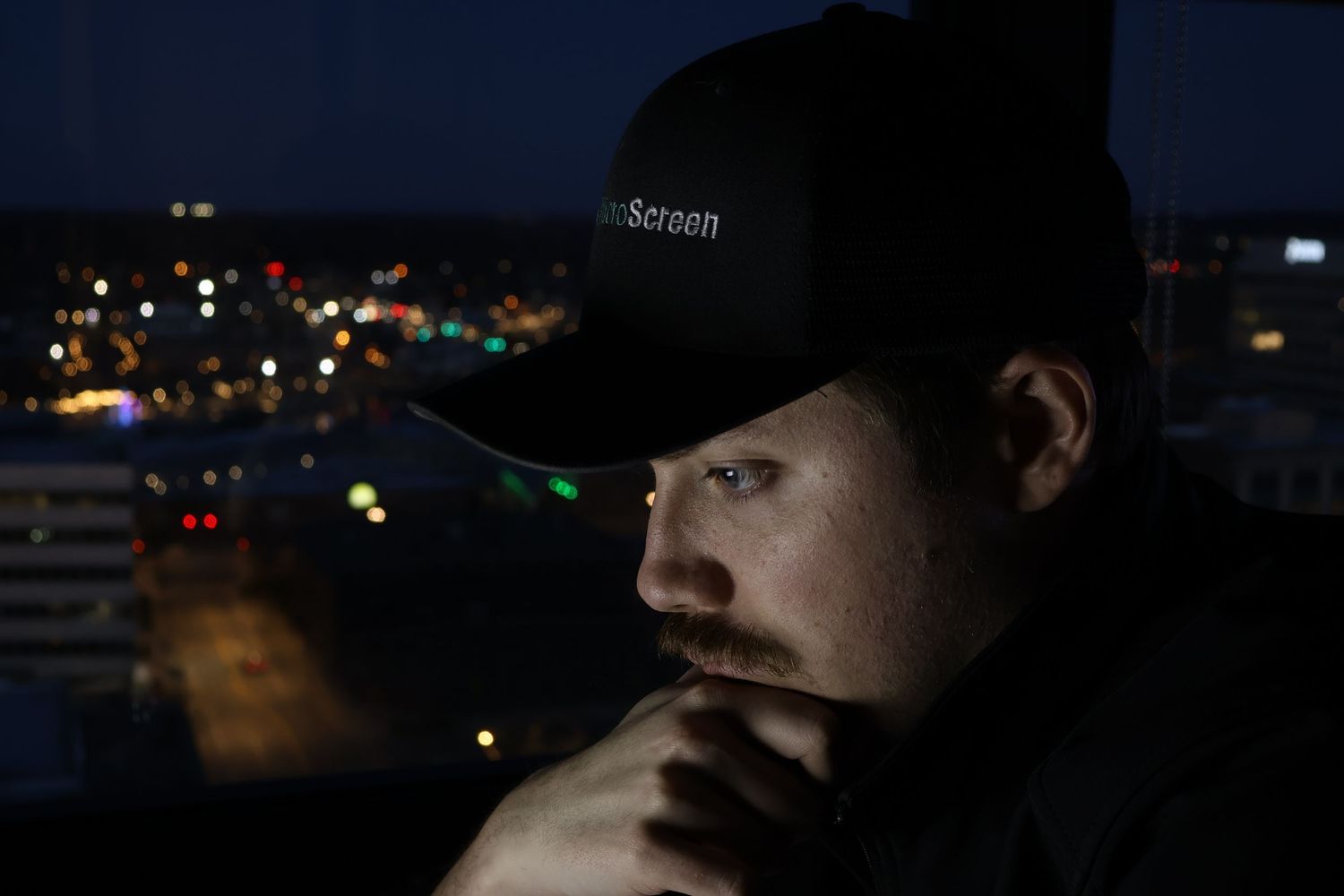 man staring at a computer in a dark room with city lights behind him