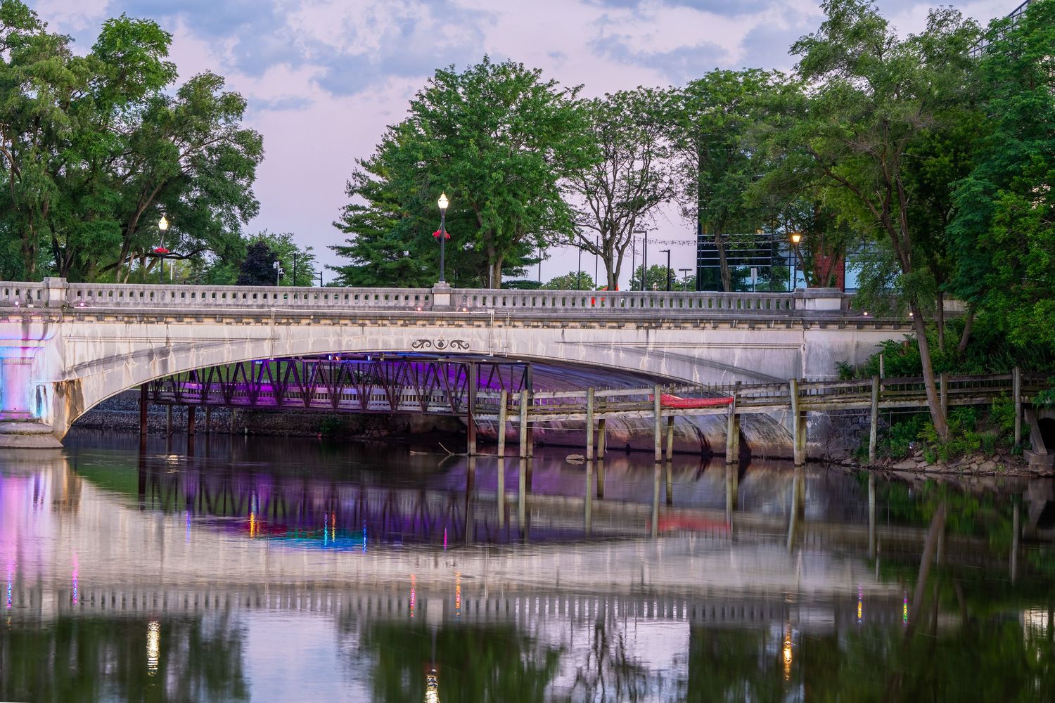 Elegant white stone bridge with purple-lit underside reflects symmetrically in calm river at dusk.