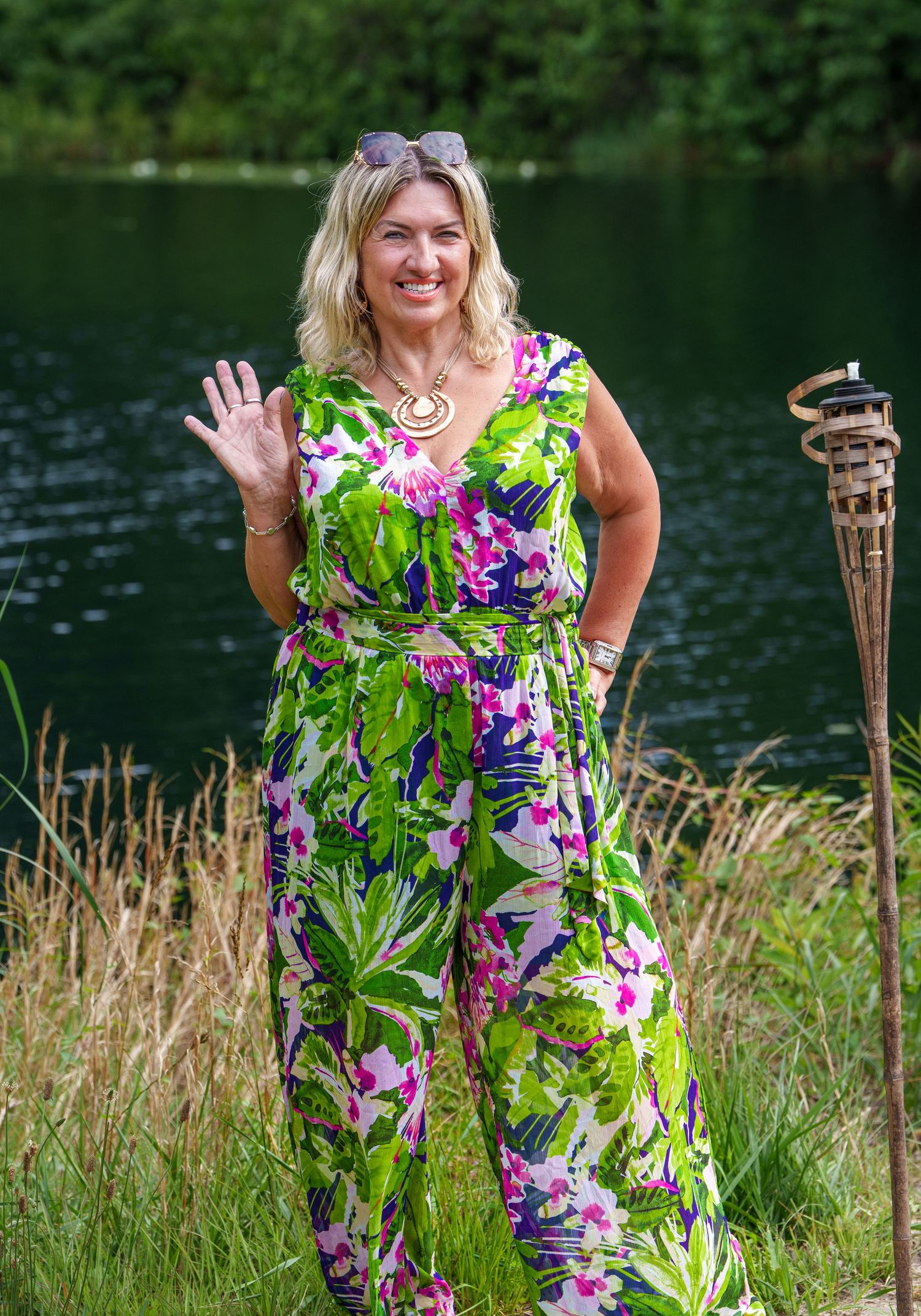 Smiling blonde woman in colorful tropical floral jumpsuit waving near a serene pond.