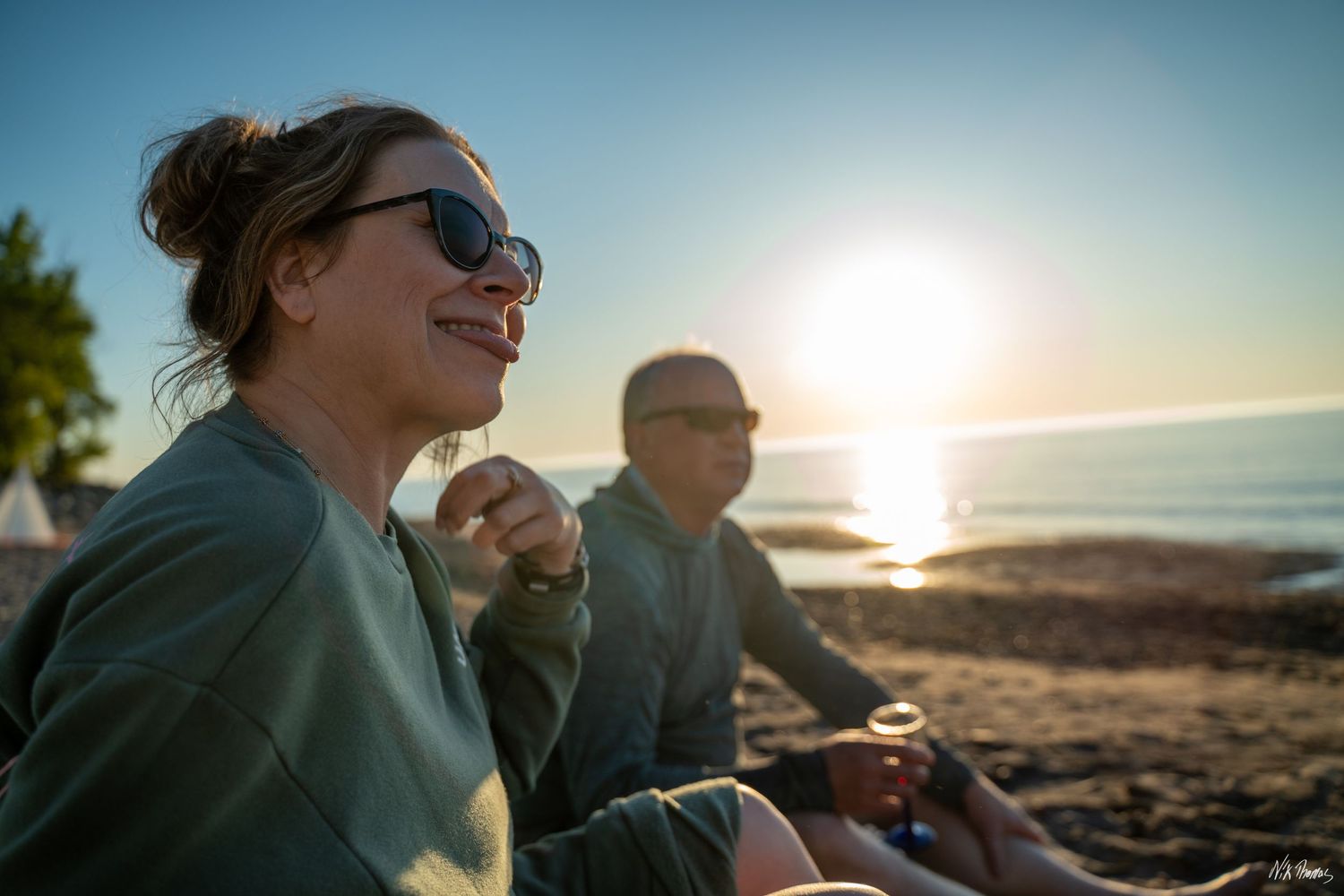 Couple smiling and relaxing on a beach at sunset, wearing green sweatshirts, with golden sunlight reflecting on calm water.