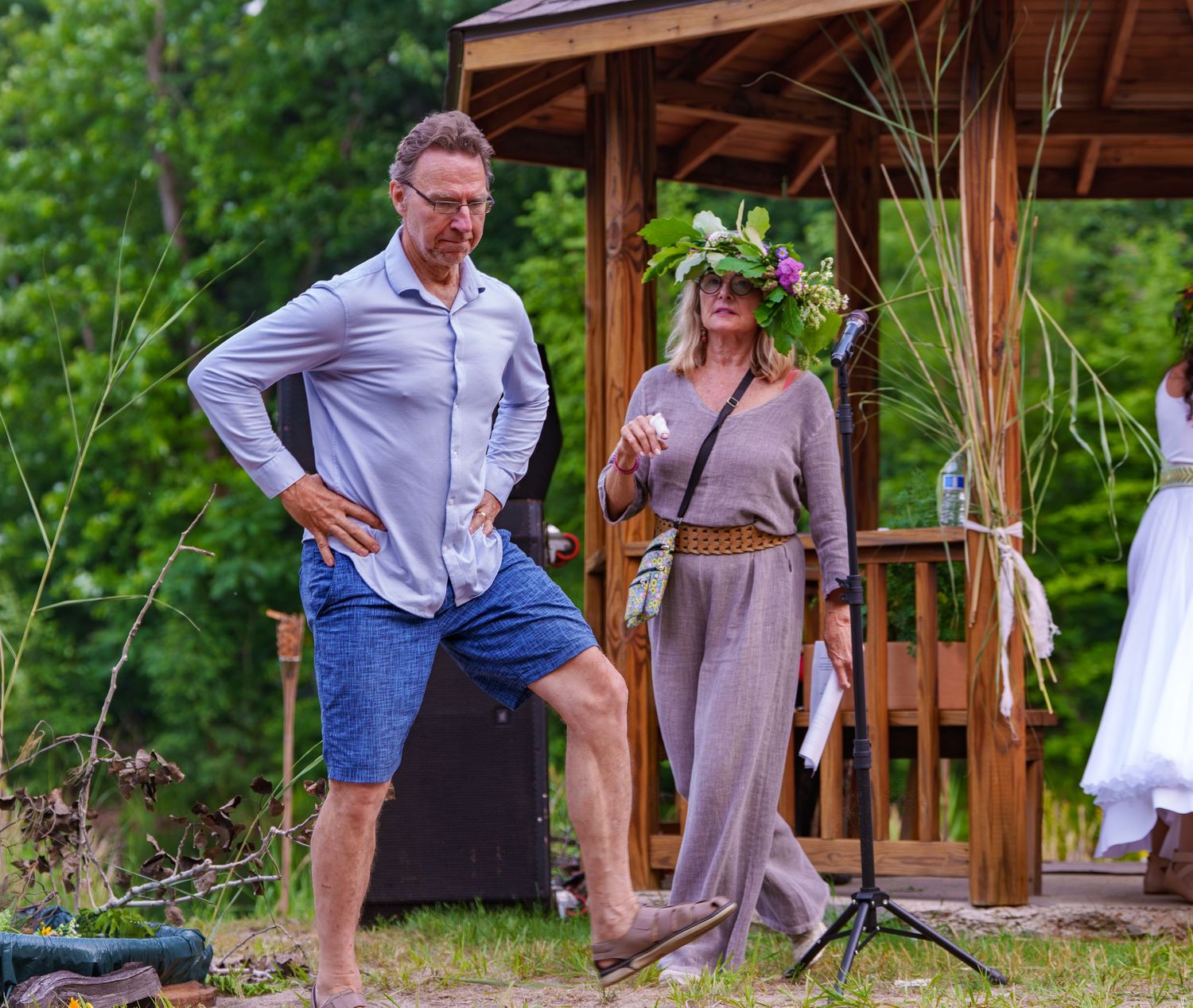 Man and woman in folk attire standing near wooden gazebo at outdoor Lithuanian cultural event.