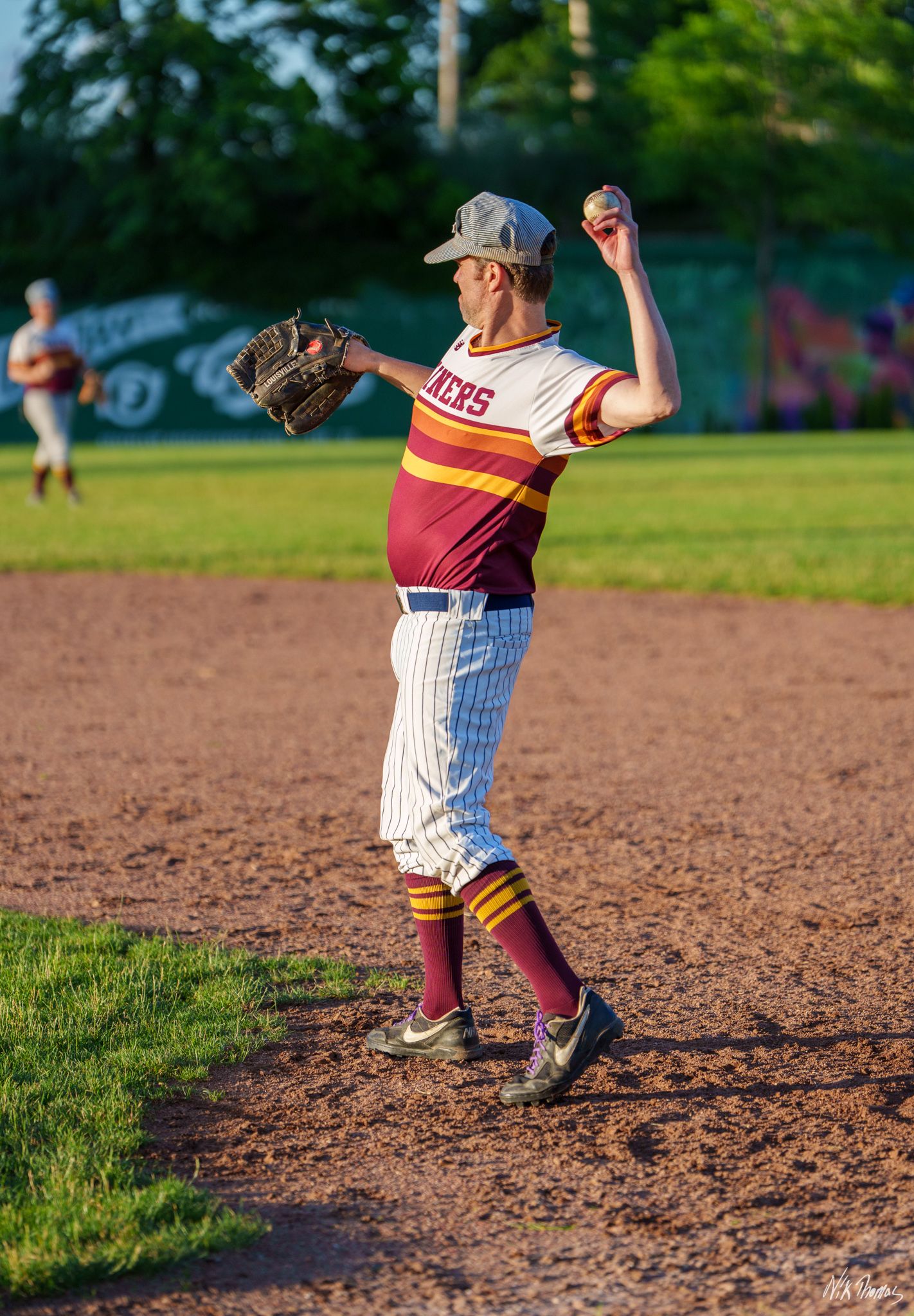 A baseball pitcher in maroon and white pinstripe uniform winds up to throw during an outdoor game.