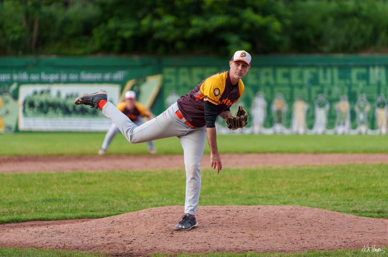 Baseball pitcher in gold and red uniform delivers a pitch during an outdoor game on a green field.