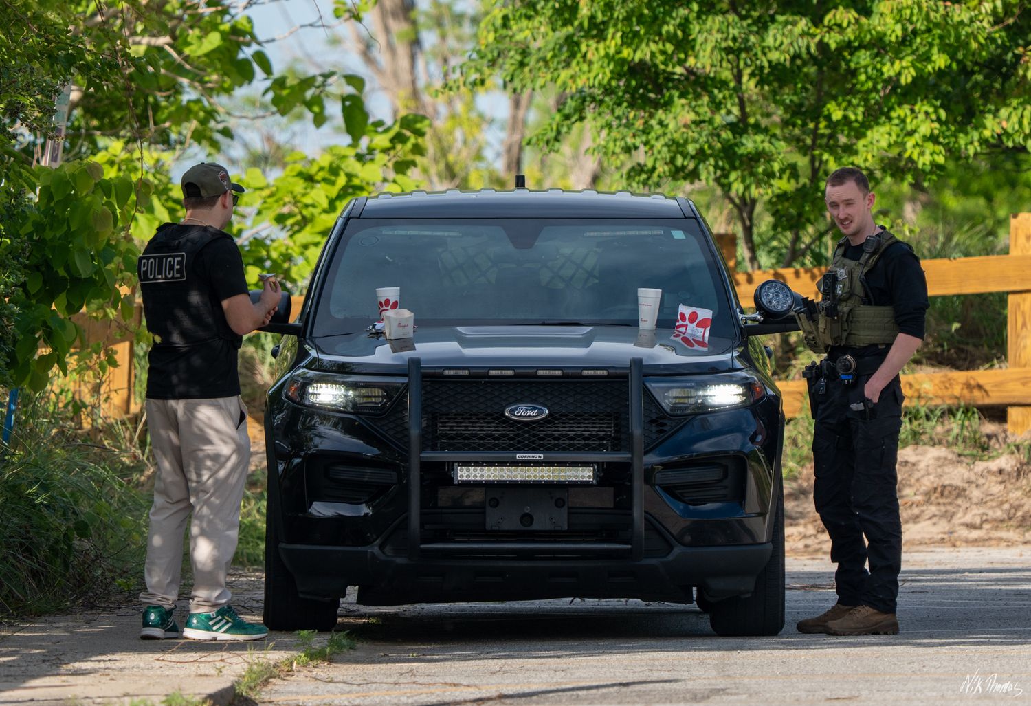 Two police officers in tactical gear stand beside a black Ford Police Interceptor SUV on a tree-lined road.