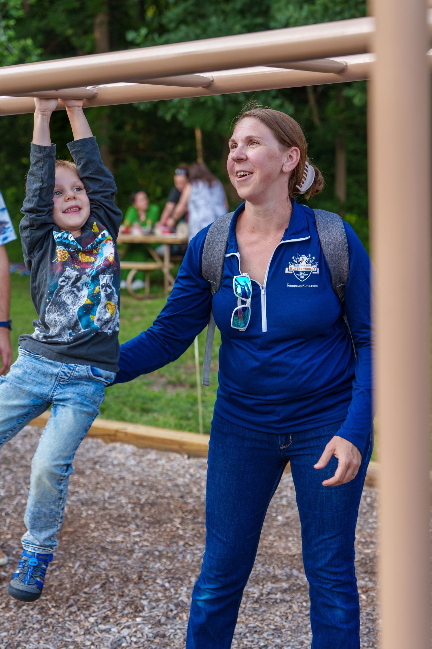 Woman in blue jacket assists a young boy playing on outdoor monkey bars at a playground.