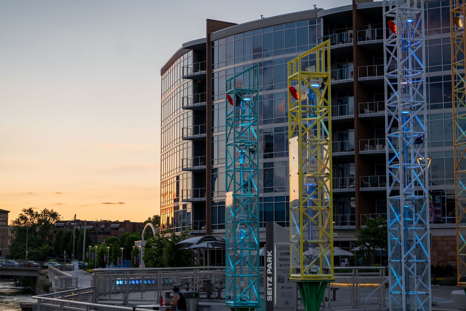 Colorful construction cranes stand beside a modern glass building at sunset near a waterfront plaza.