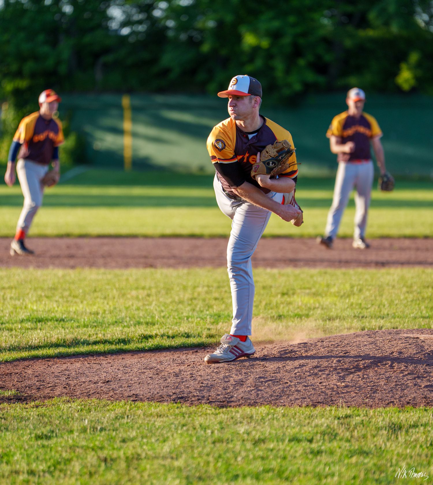 Baseball pitcher in yellow uniform mid-throw on mound during evening game with teammates visible in background.