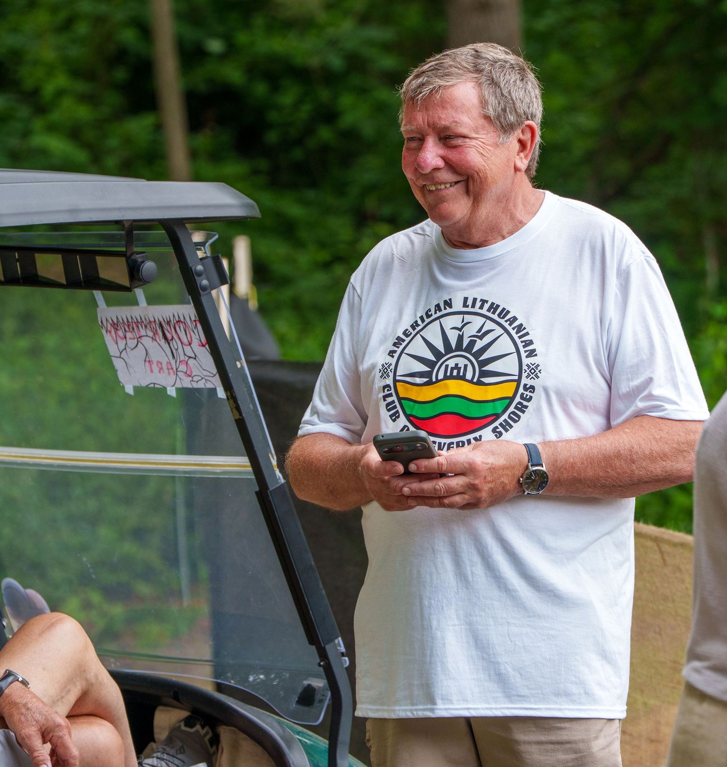 Smiling man in American Lithuanian Club of Beverly Shores t-shirt holding phone near golf cart.