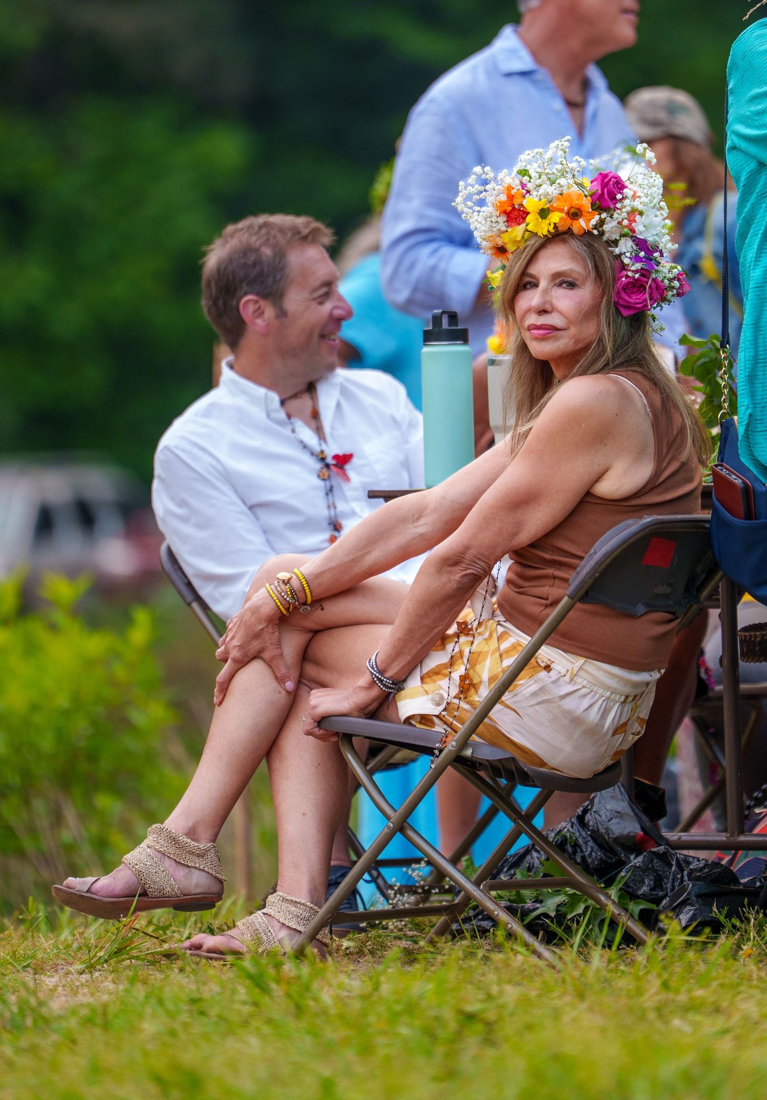 Woman wearing colorful flower crown sits in folding chair outdoors at a summer festival event.