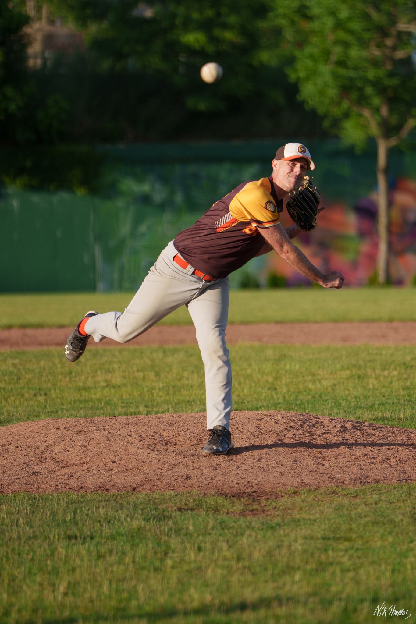 Baseball pitcher in maroon and gold uniform mid-throw on mound during evening game on green field.
