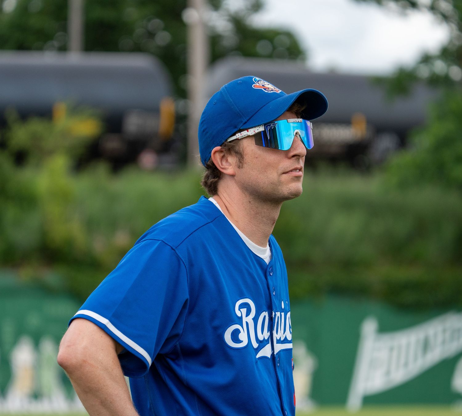 Baseball player in blue Raptors jersey and mirrored sunglasses stands alert on field during game.