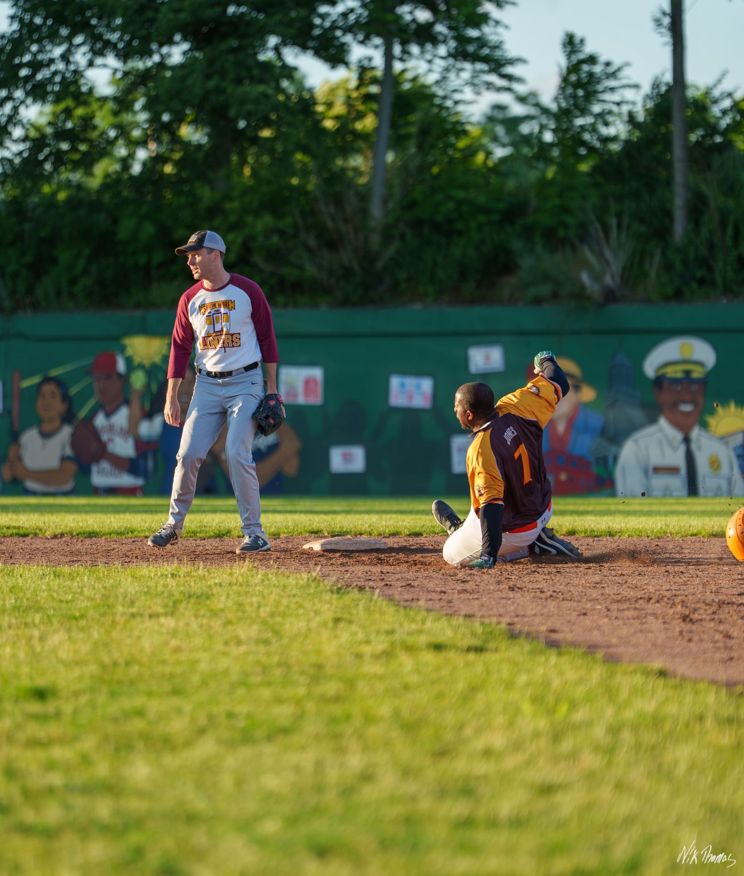 A baseball player slides into second base as the fielder stands nearby during a sunny evening game.