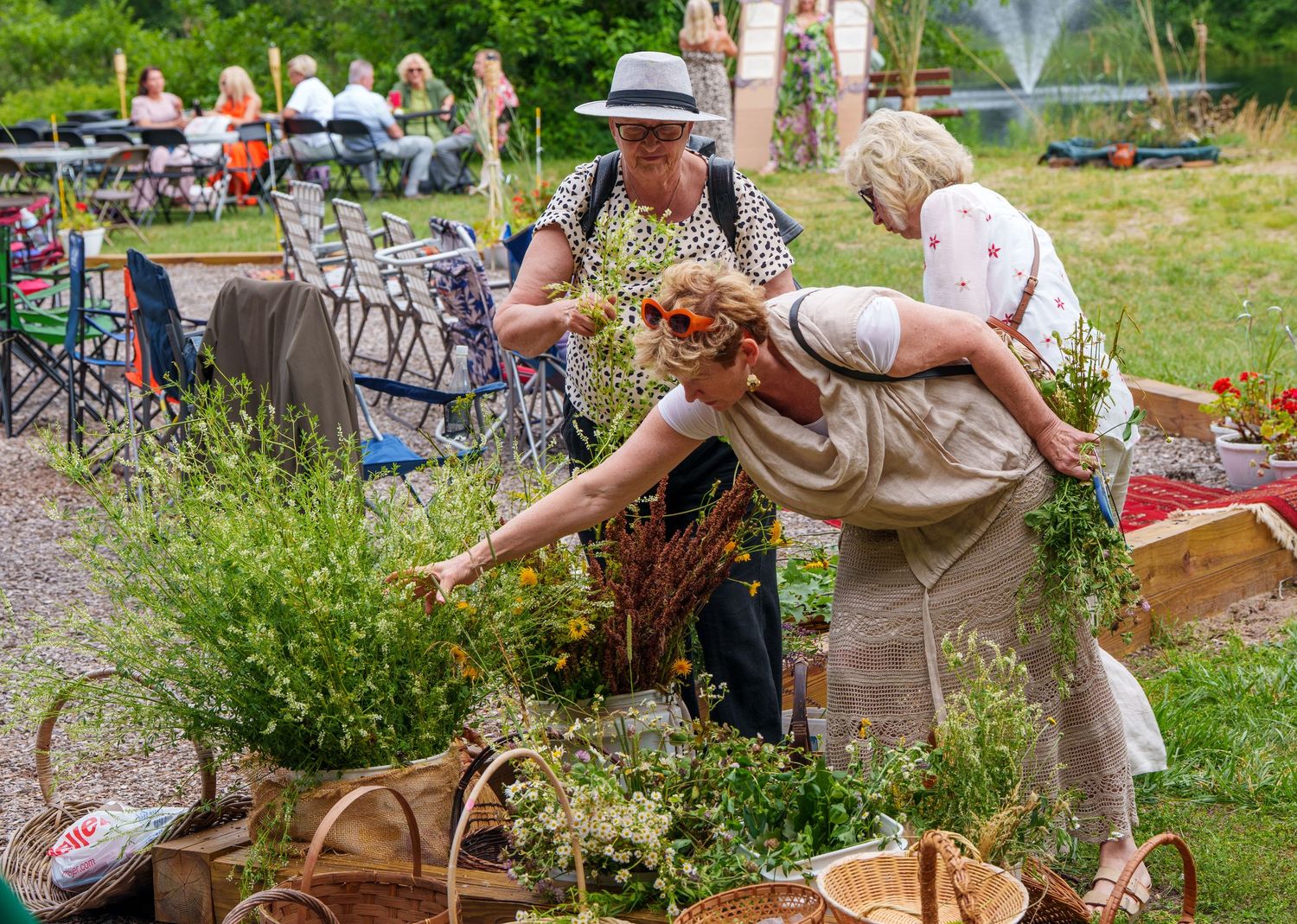 Women arranging wildflowers and herbs in wicker baskets at an outdoor midsummer festival celebration.