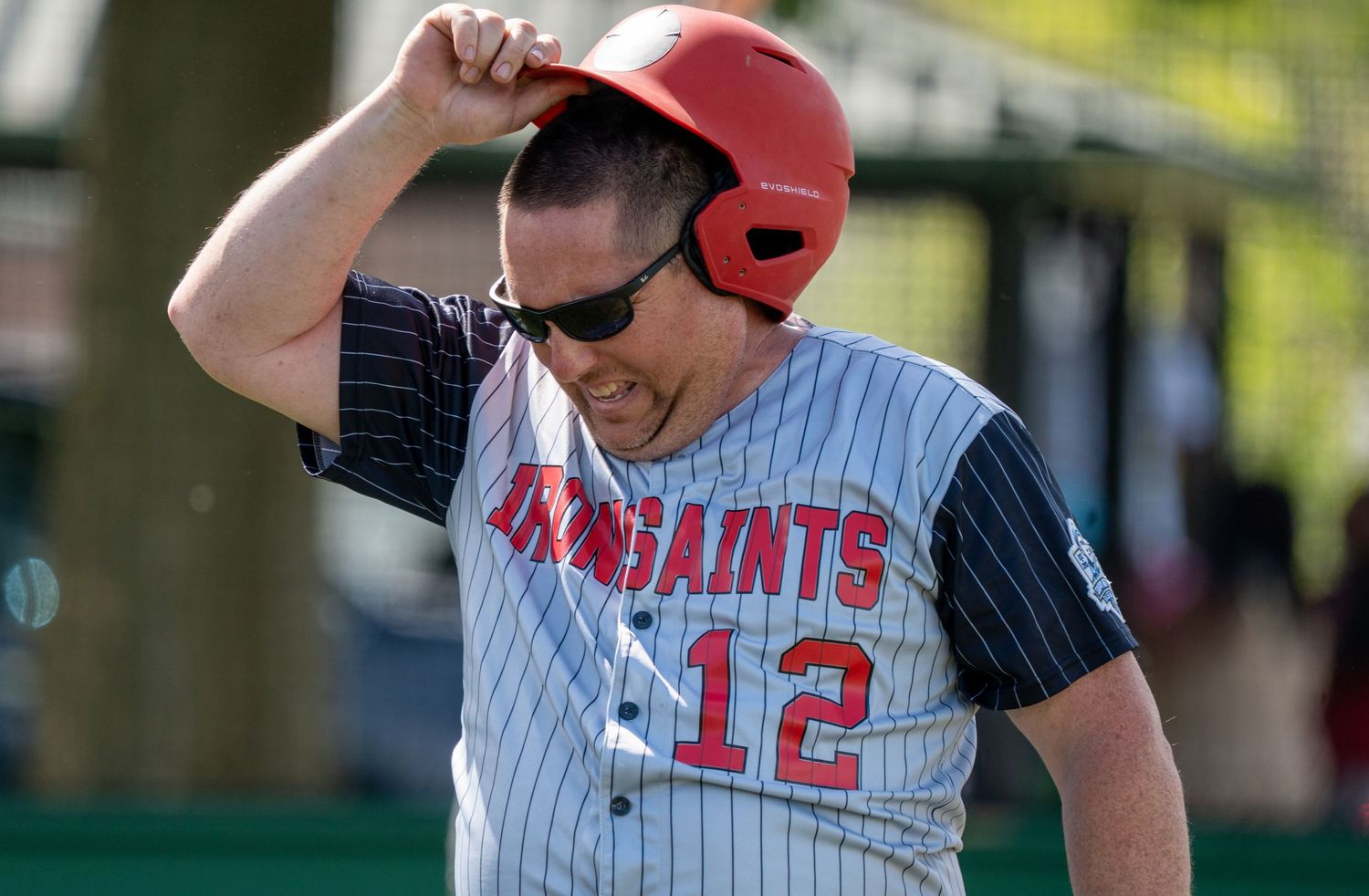 Baseball player #12 in red helmet and GoSaints uniform adjusts helmet on field during game.
