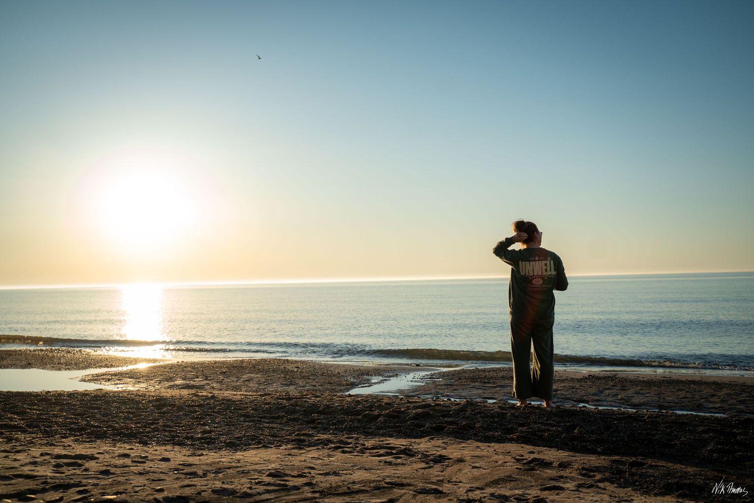 Person standing on a sandy beach at sunset, gazing at the horizon over calm ocean waters in warm golden light.