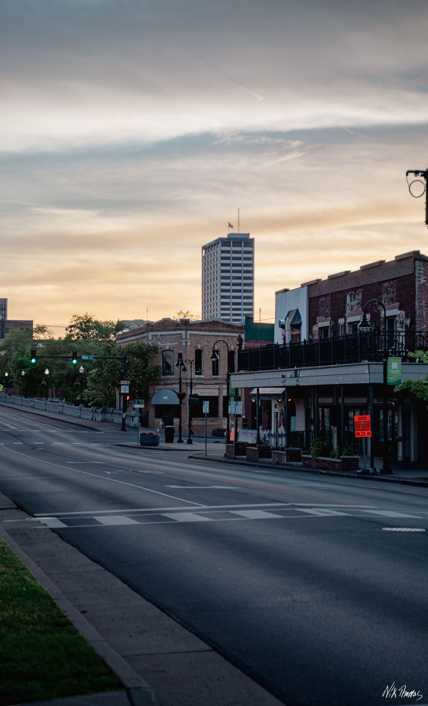 Empty urban street at sunset with brick storefronts, green traffic lights, and a tall skyscraper against a golden sky.
