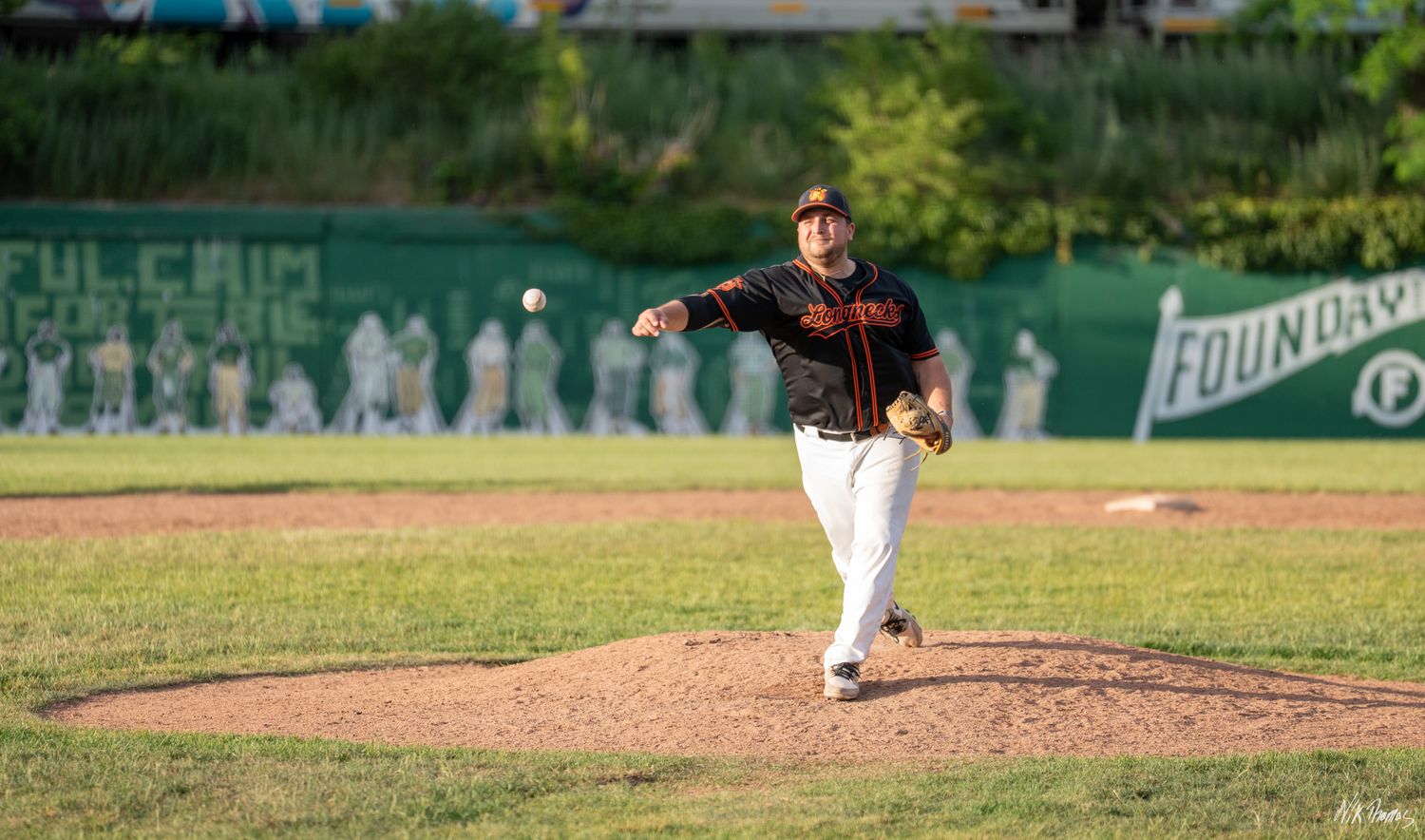 Baseball pitcher in black and orange uniform mid-throw on a sunny green field during a game.