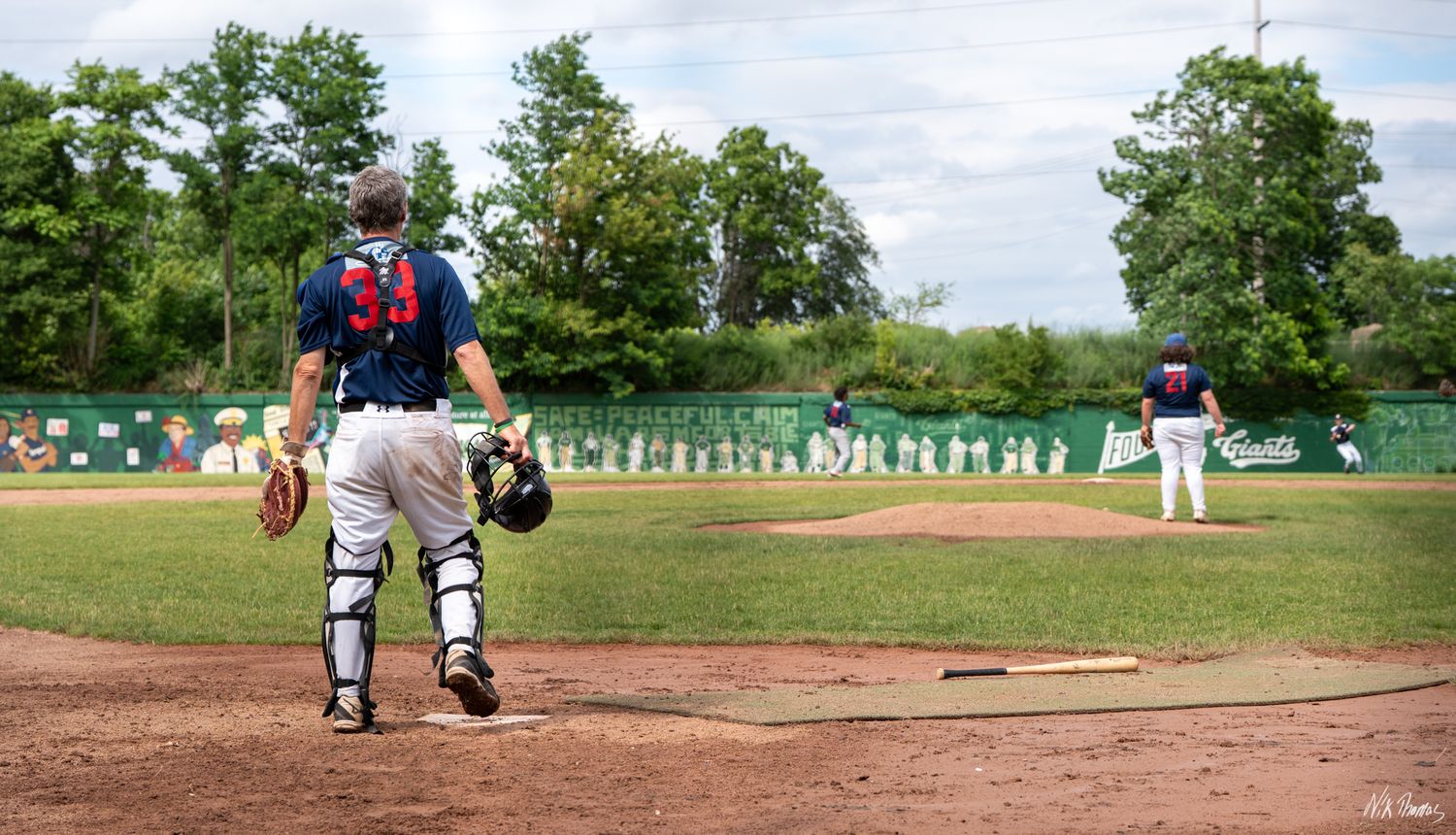 Baseball catcher in full gear watches pitcher on mound during a game with dugout visible in background.