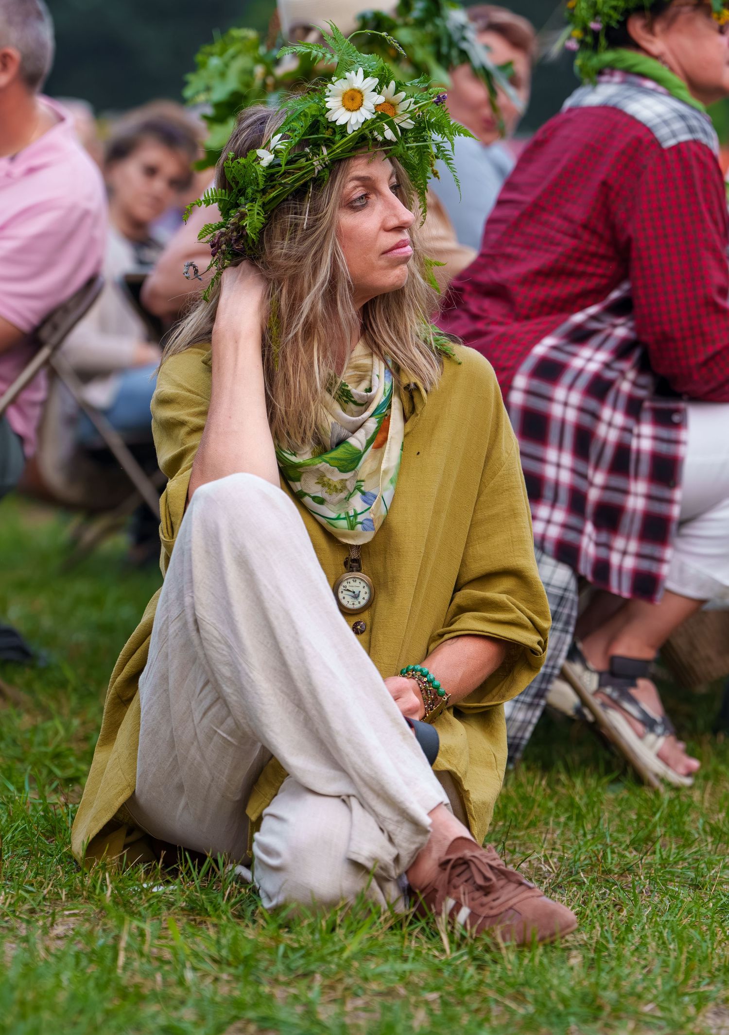 Woman wearing a floral daisy crown sitting cross-legged on grass at an outdoor summer festival.