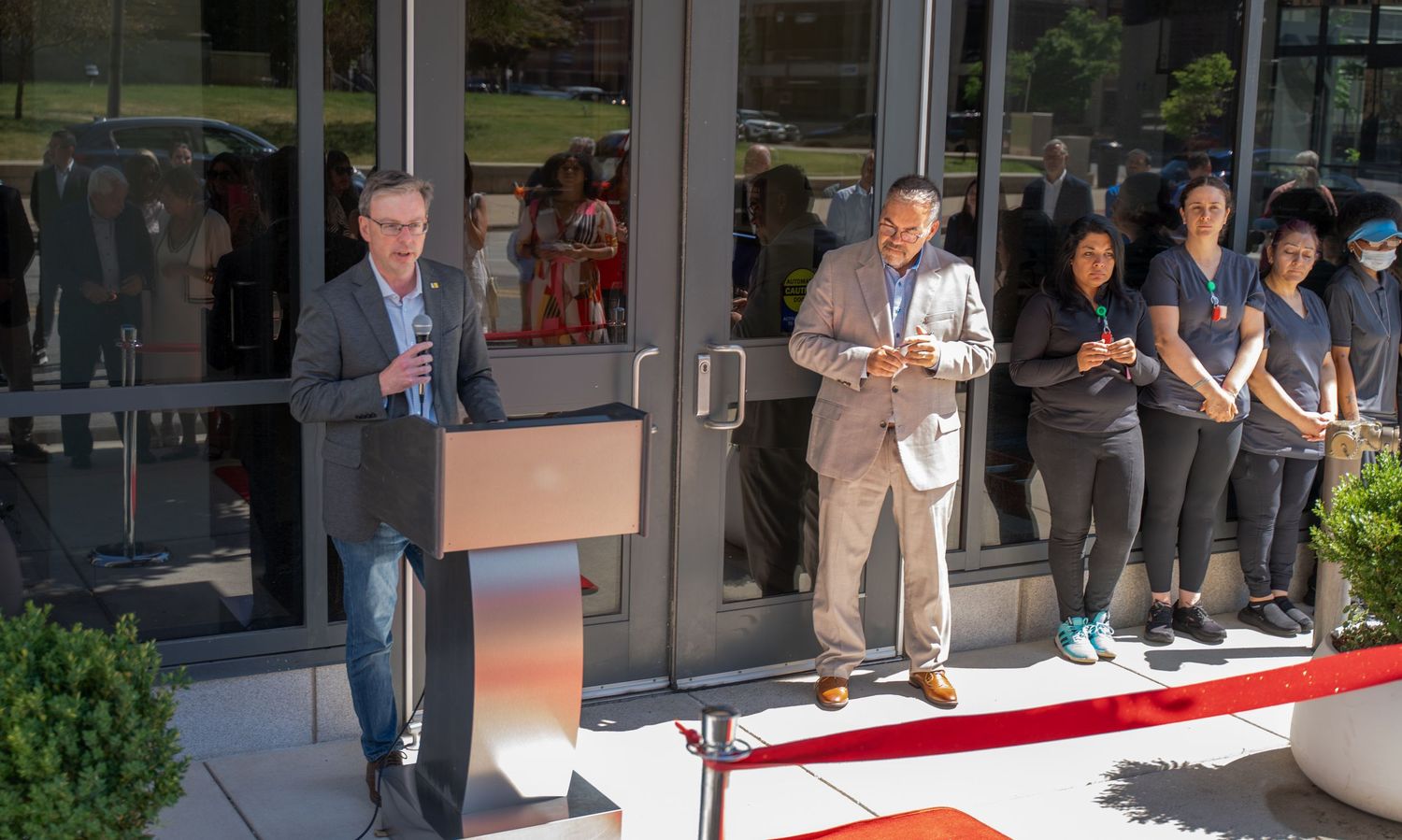 Two men speak at a podium during a ribbon-cutting ceremony outside a modern building with staff lined up.