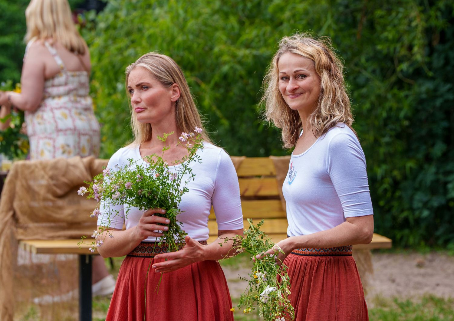 Two blonde women in white tops and red skirts holding flower bouquets at an outdoor festival.