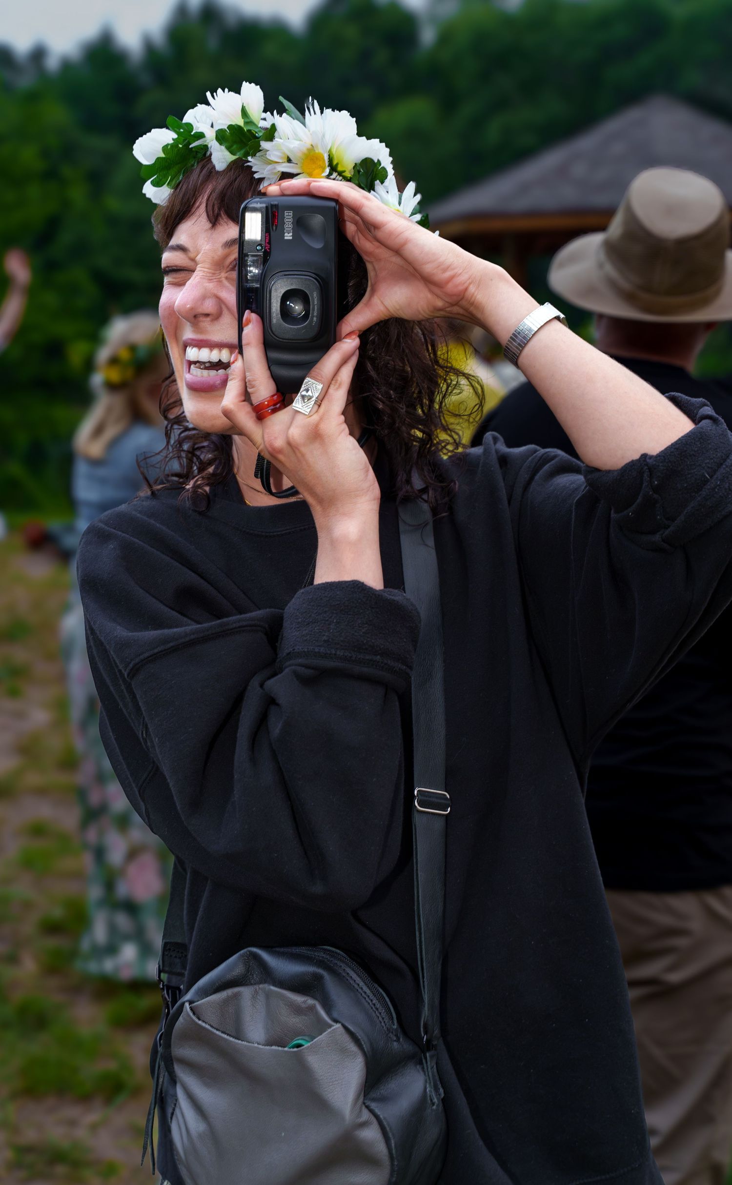 Smiling woman wearing a white flower crown takes a photo with an instant camera at an outdoor festival.