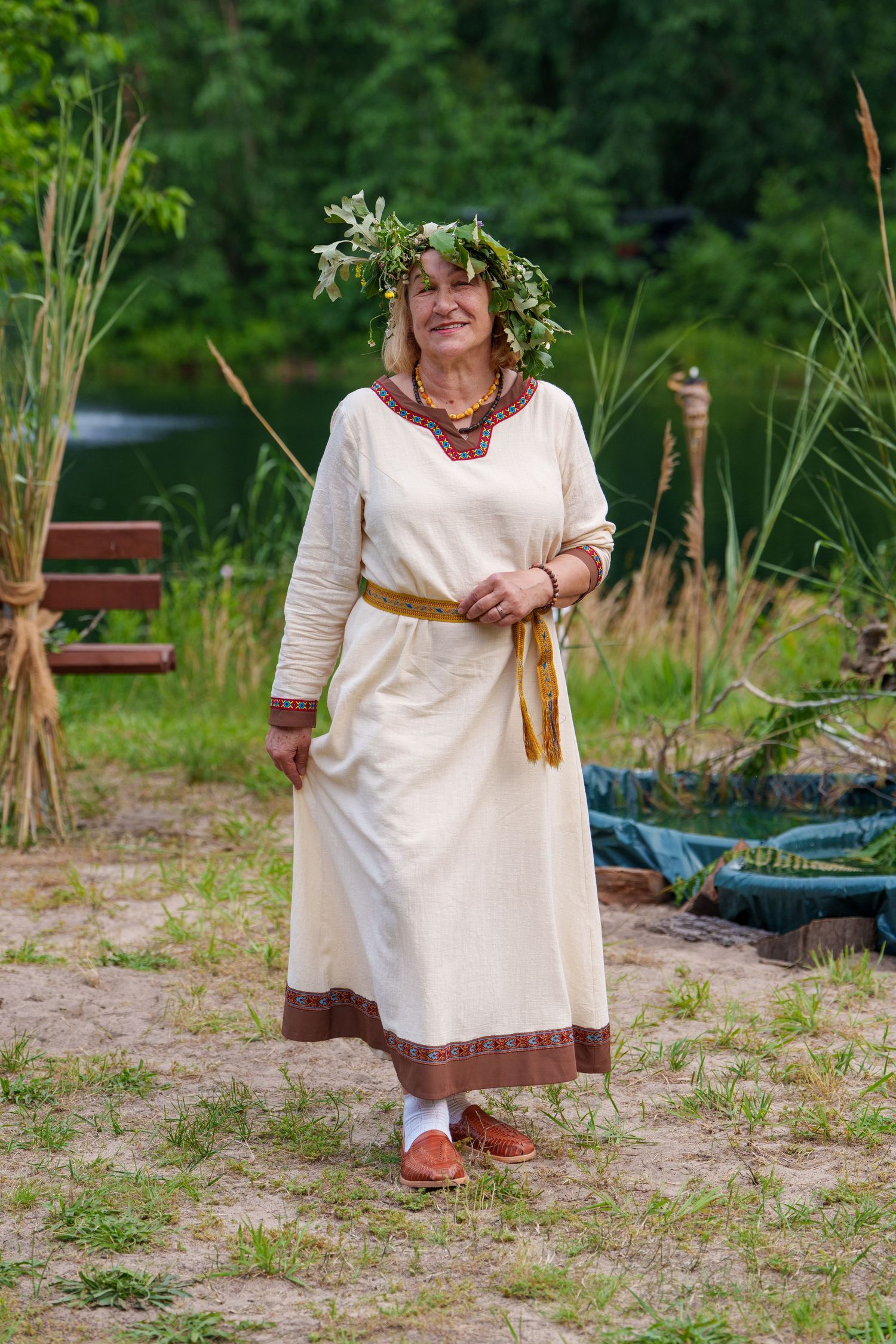 Woman in traditional Eastern European folk costume with floral wreath crown standing near a river in a green forest setting.
