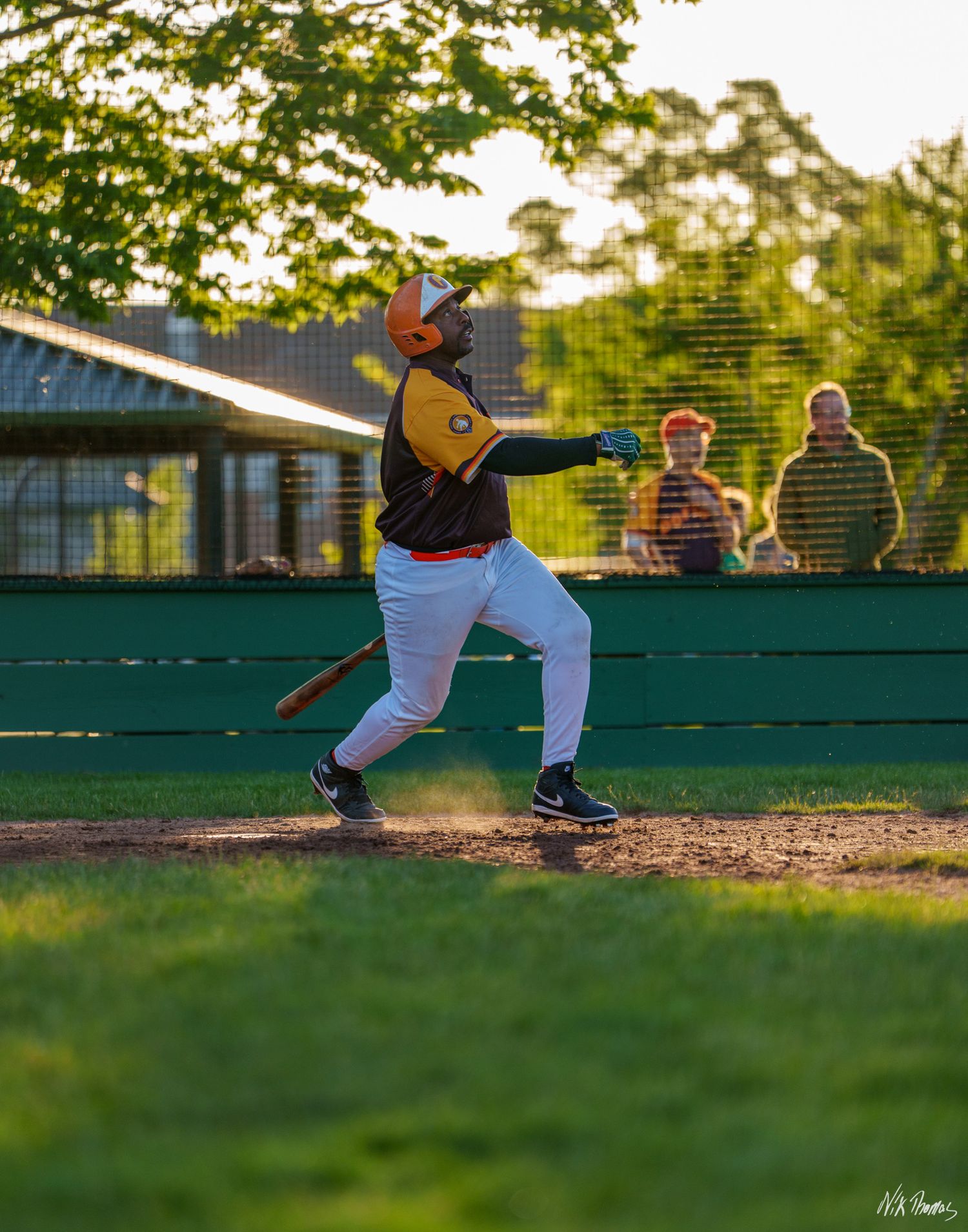 Baseball batter swinging at pitch during evening game with spectators watching from behind green outfield fence.