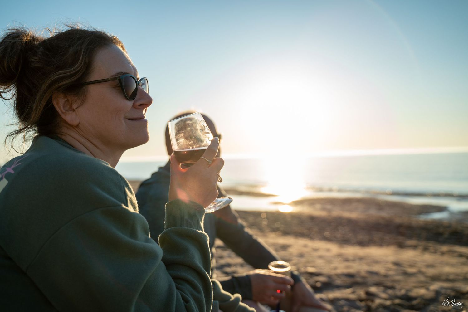 Woman enjoying wine on a beach at sunset, wearing sunglasses and a green sweatshirt, with ocean views.