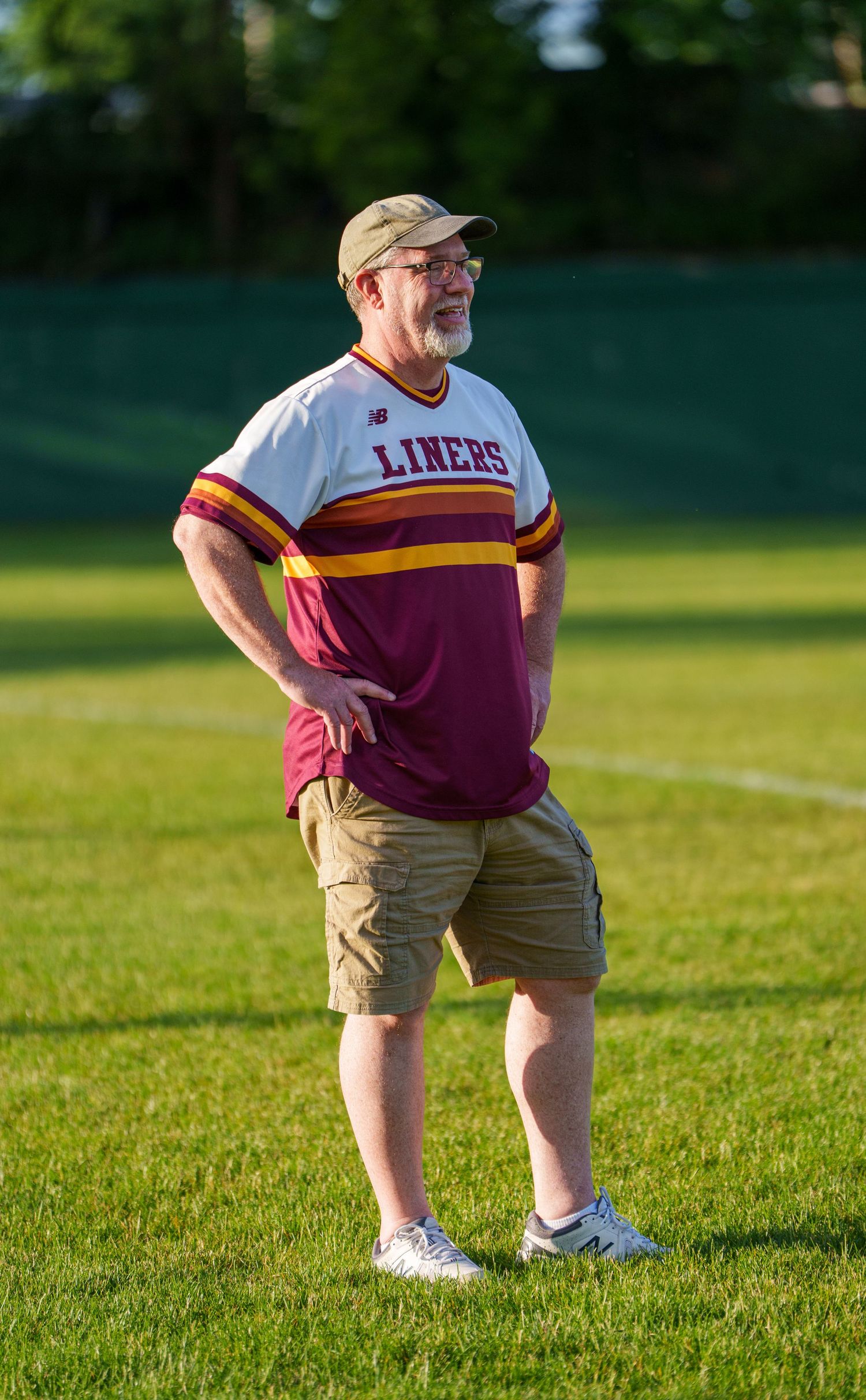 Man wearing a Liners baseball jersey and khaki shorts standing on a green sports field outdoors.