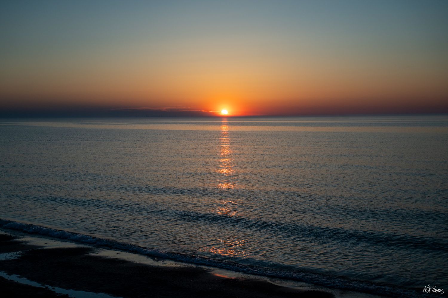 A stunning ocean sunset with vibrant orange and red hues reflecting on calm waves along a serene beach shoreline.