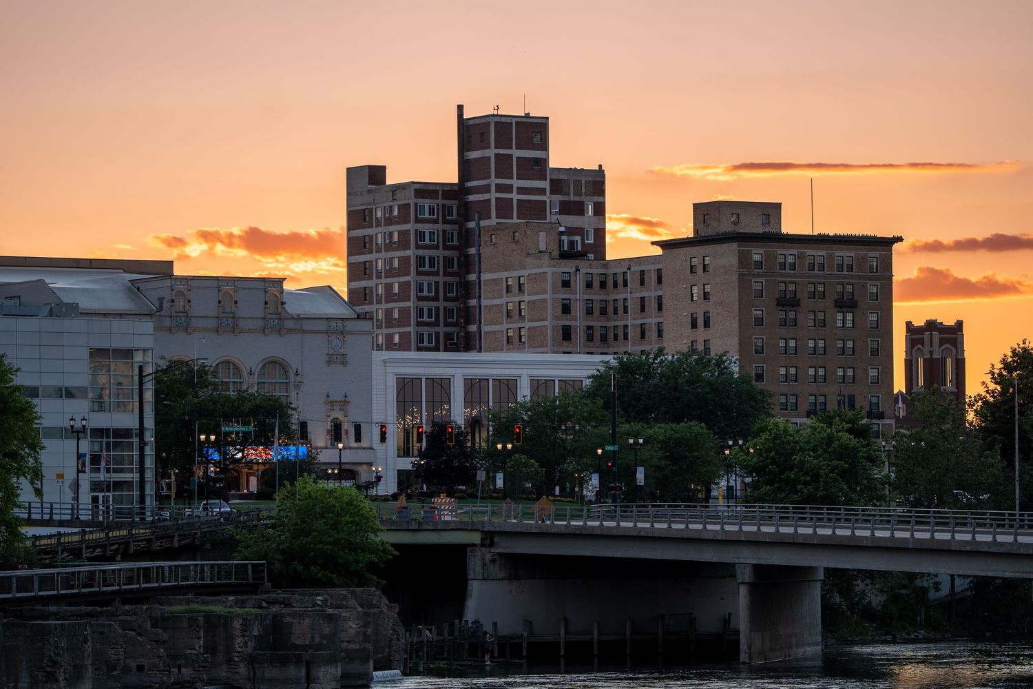 Sunset skyline over a river city with brick buildings and a bridge glowing under an orange dusk sky.