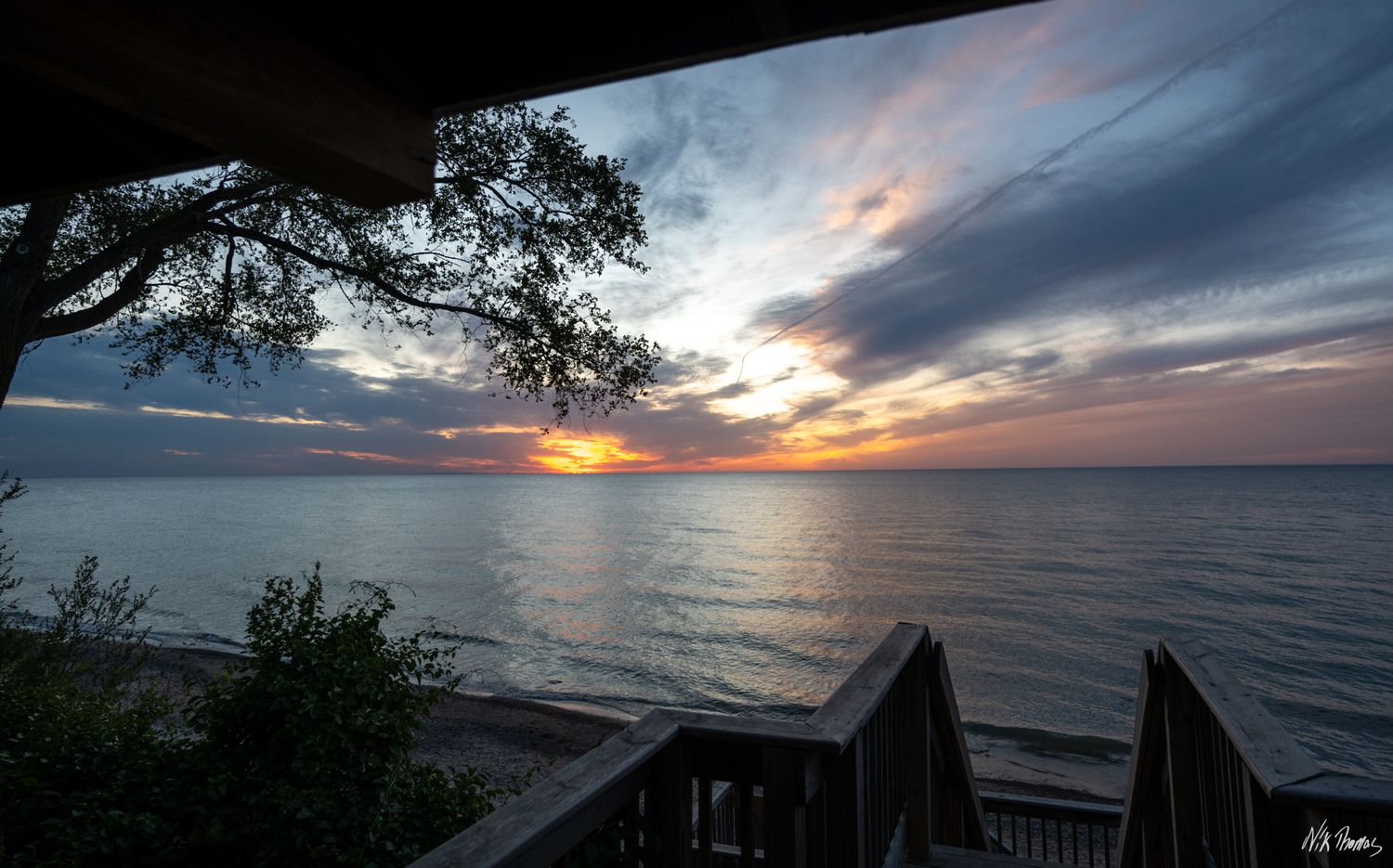 A breathtaking lakeside sunset viewed from a wooden deck with a silhouetted tree and colorful cloudy sky.