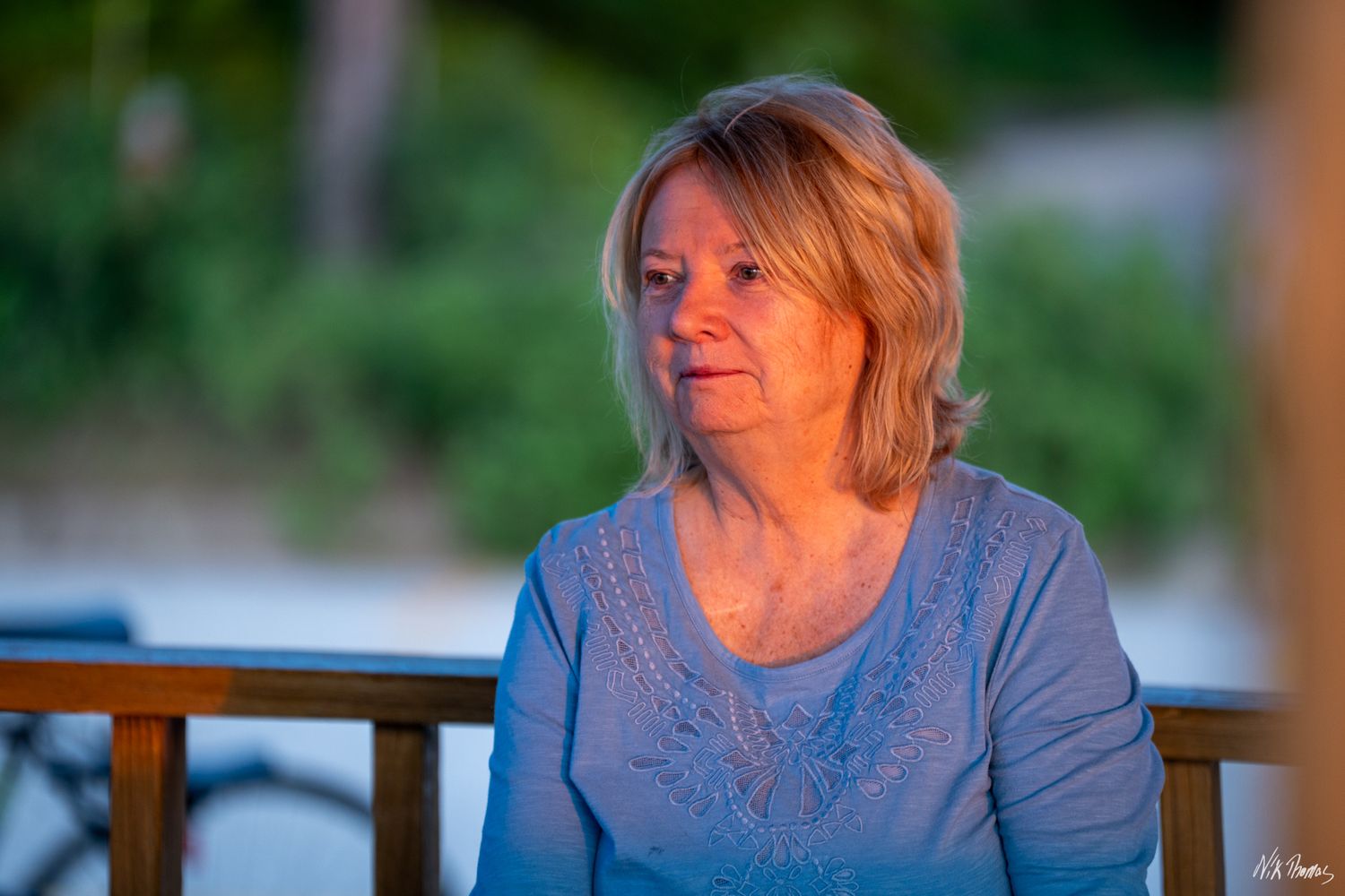 Older blonde woman in blue top sitting outdoors at dusk, bathed in warm golden light with serene expression.