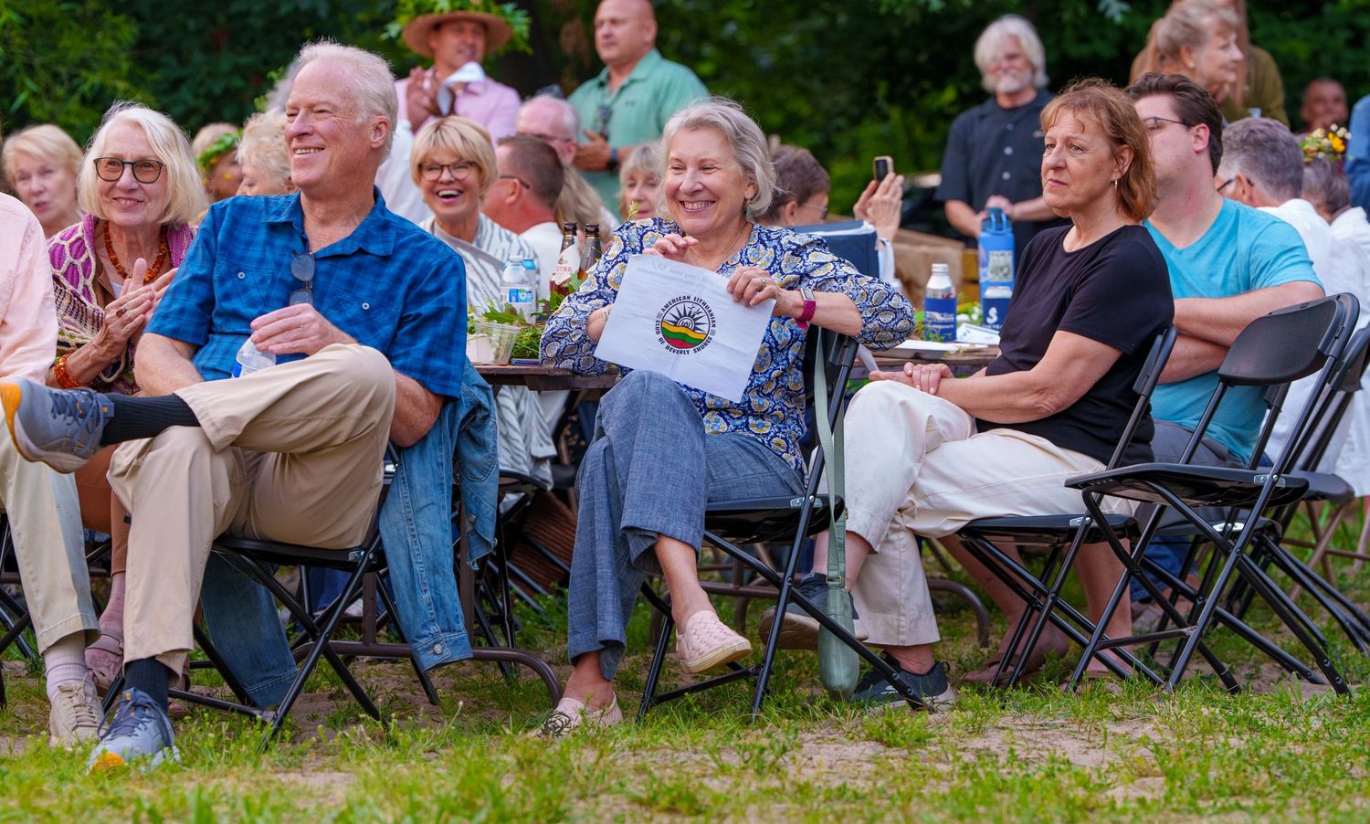 Group of smiling adults enjoy outdoor summer event seated on folding chairs.