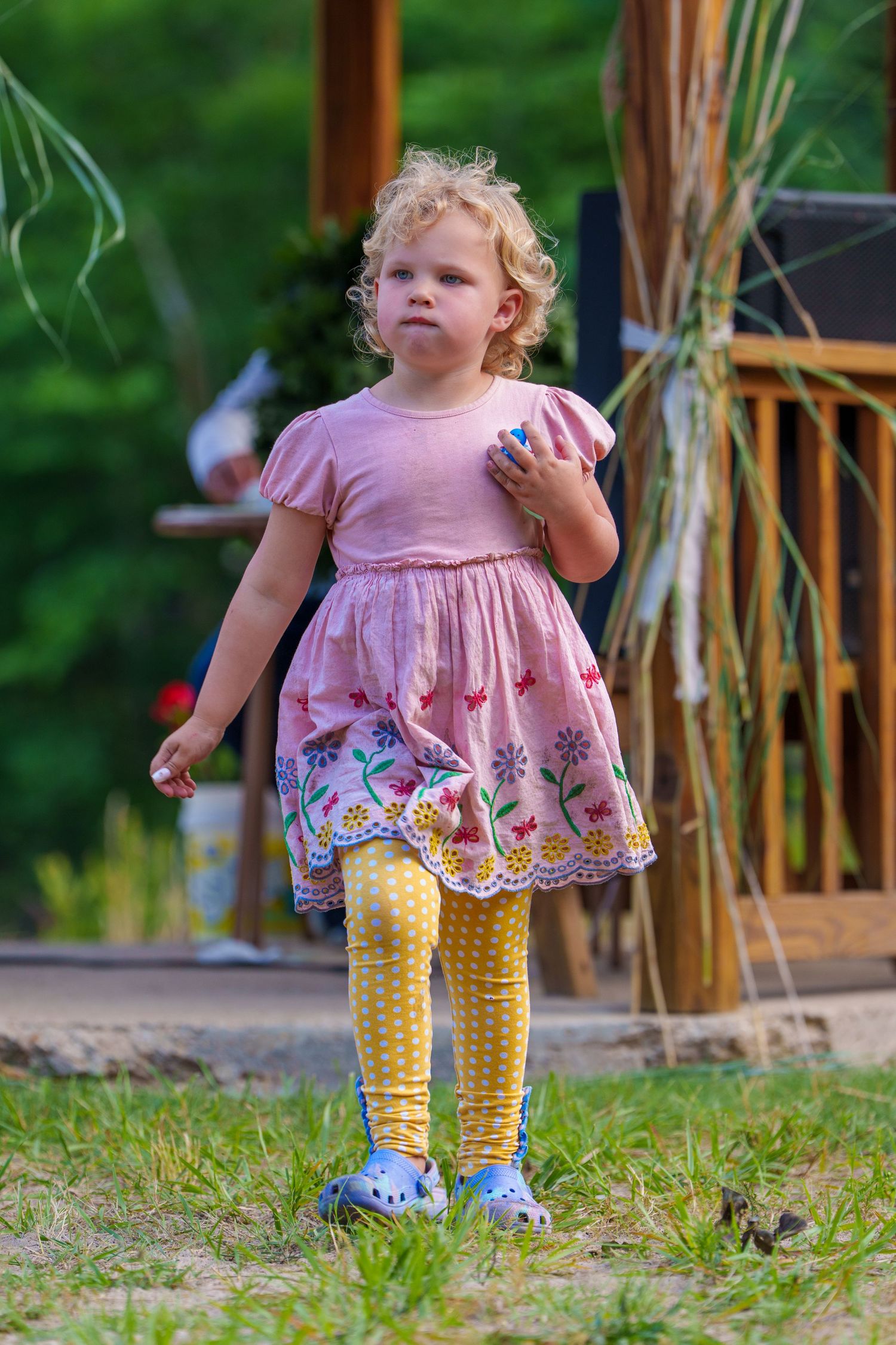 Curly-haired toddler girl in pink floral dress and yellow leggings playing outdoors in a garden.