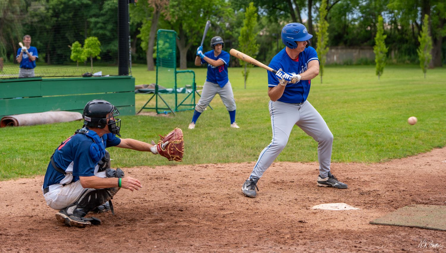 Baseball batter in blue uniform preparing to hit pitch with catcher and players in background.