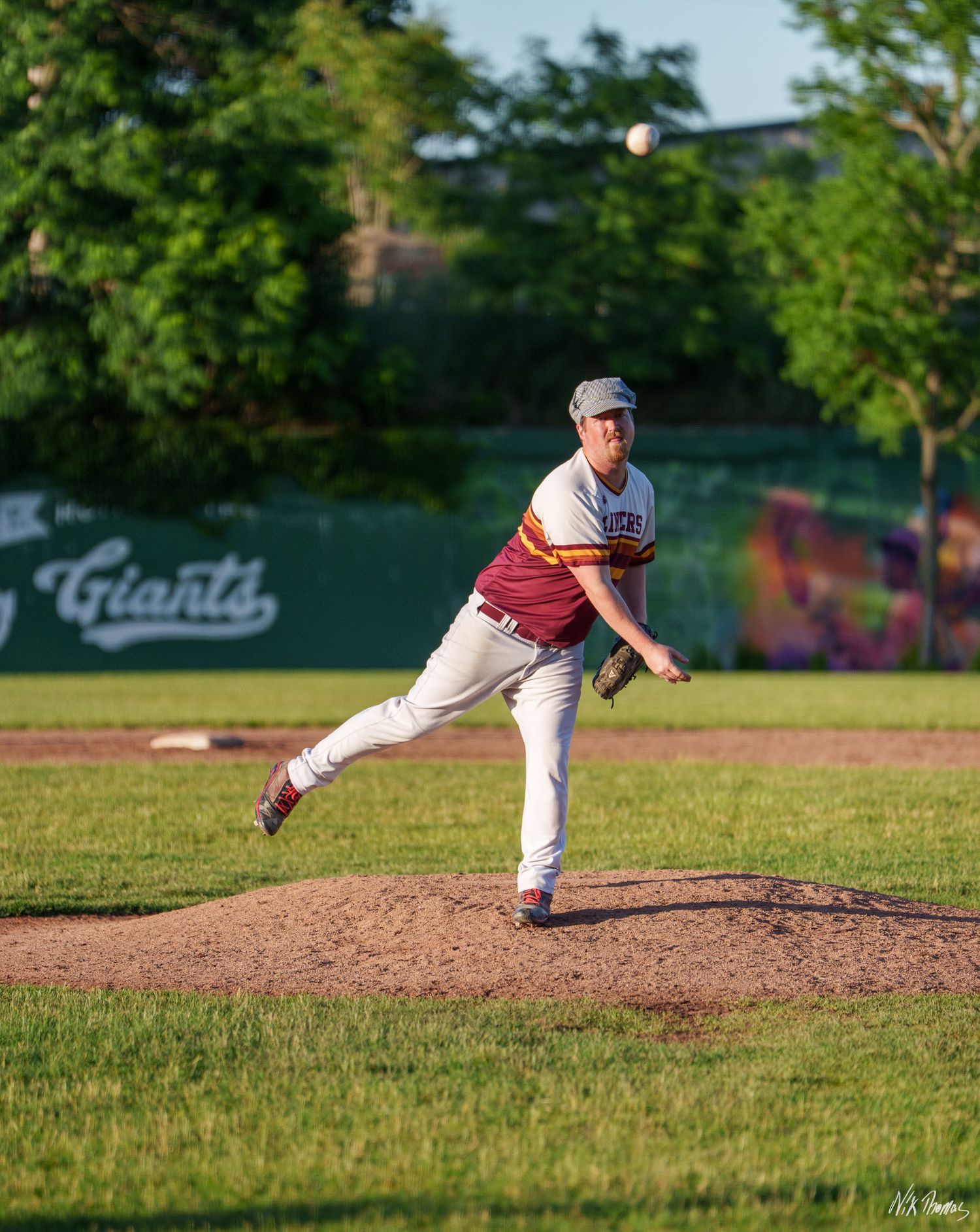 Baseball pitcher in maroon and white uniform mid-throw on a green field near a Giants dugout.