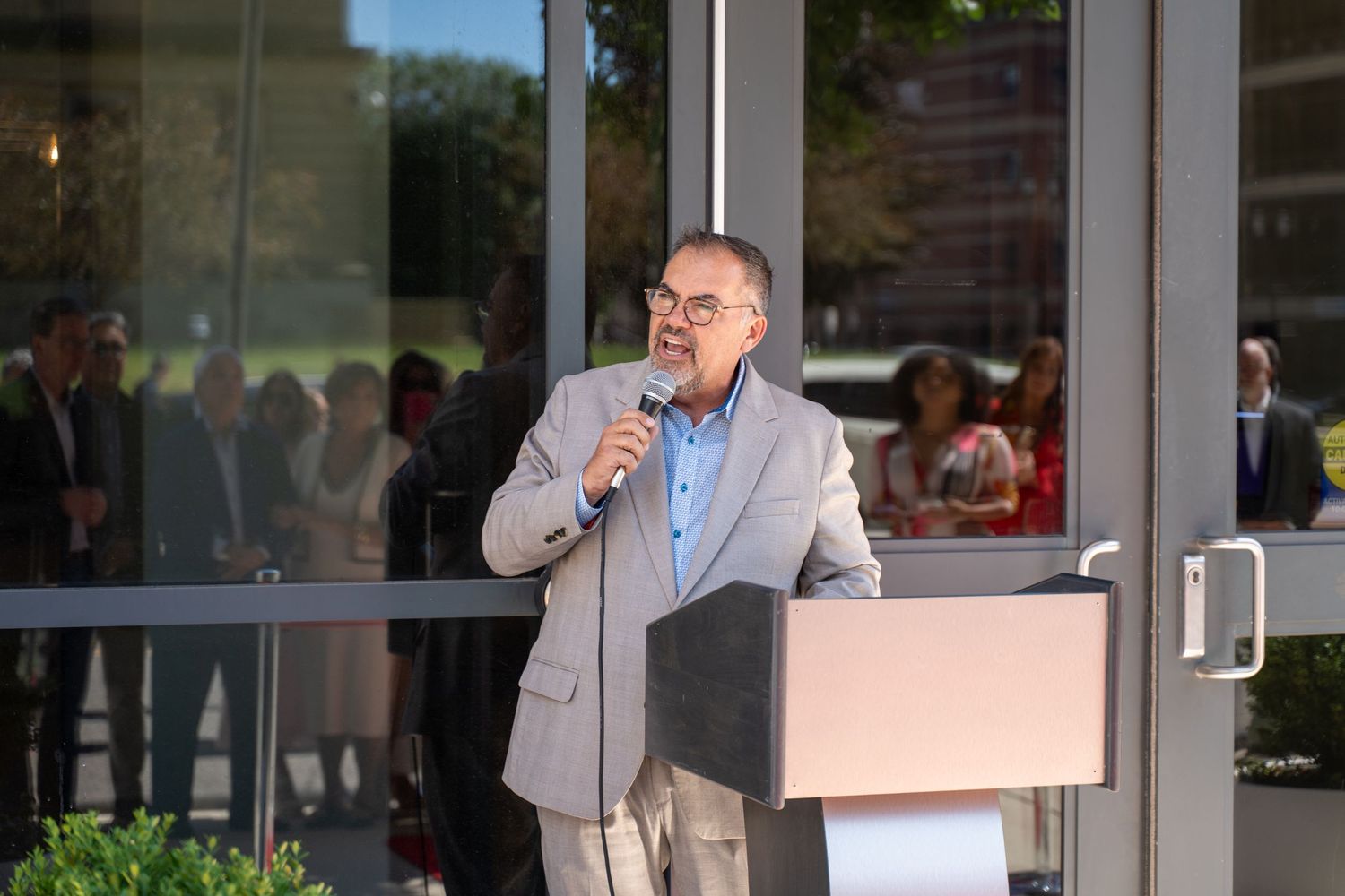 Man in gray blazer speaking at podium with microphone at outdoor corporate event with crowd in background.