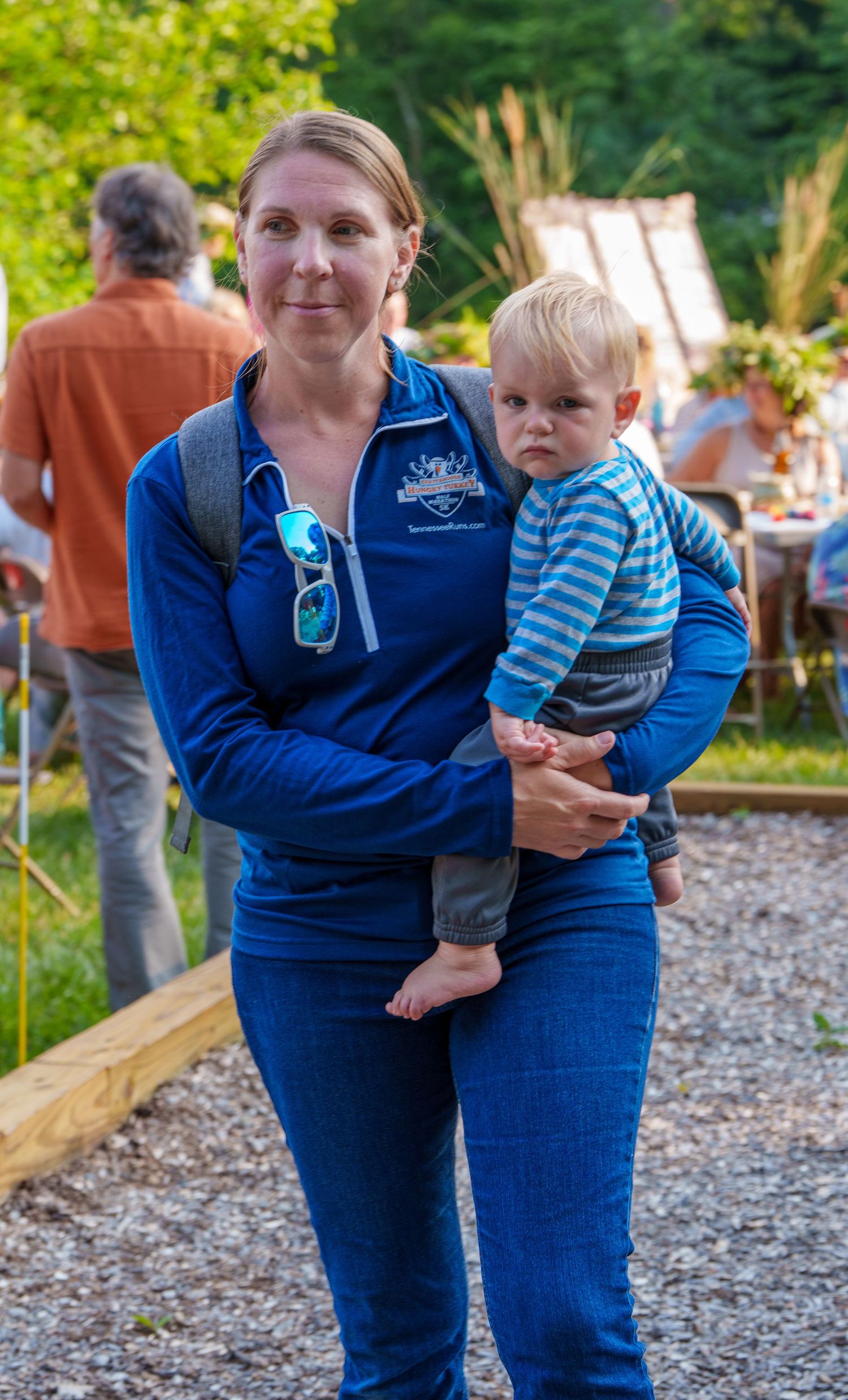 Woman in blue jacket carrying a barefoot toddler boy at an outdoor summer event.