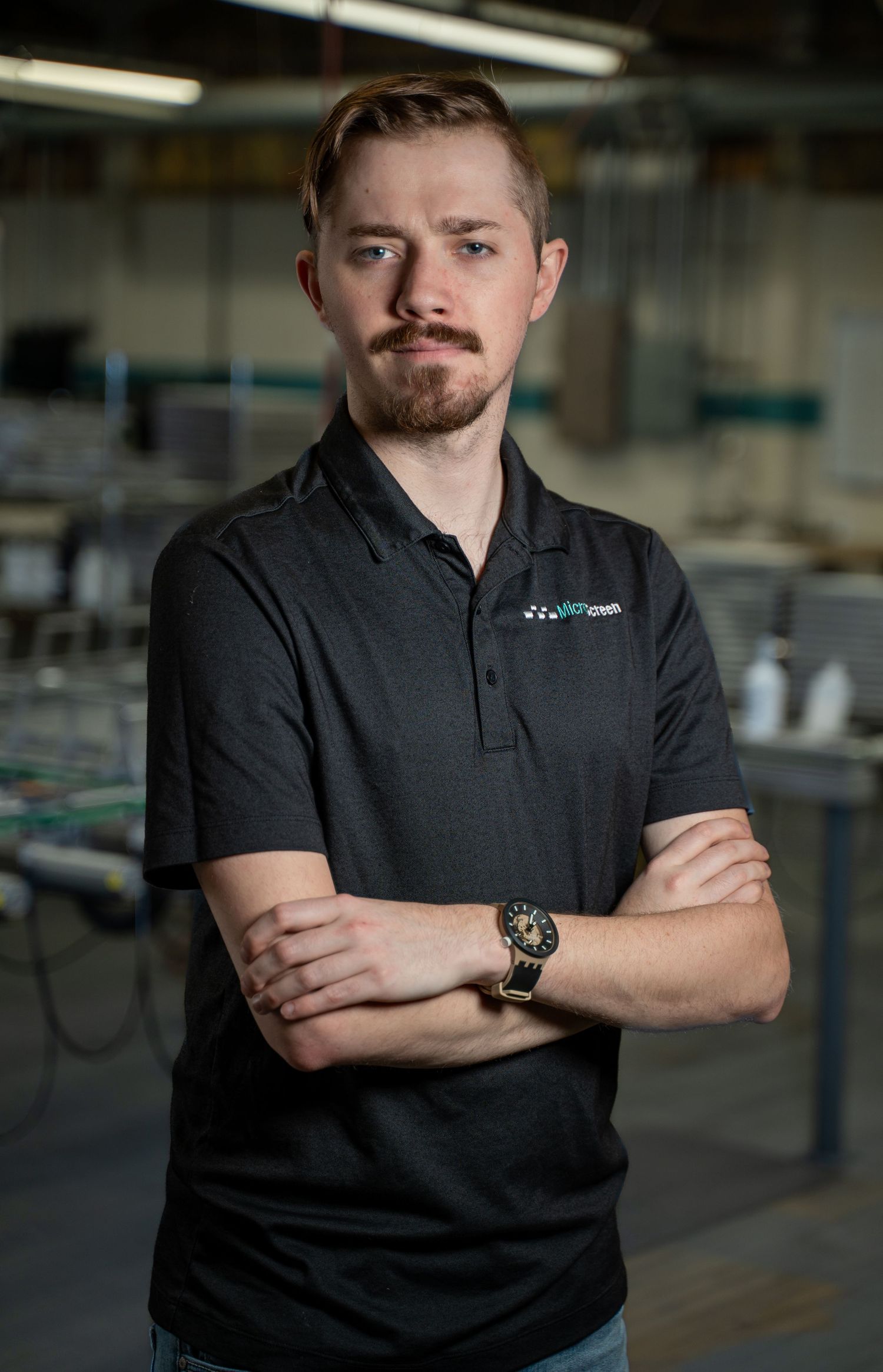 Young man with beard in black polo shirt stands with arms crossed in an industrial warehouse setting.